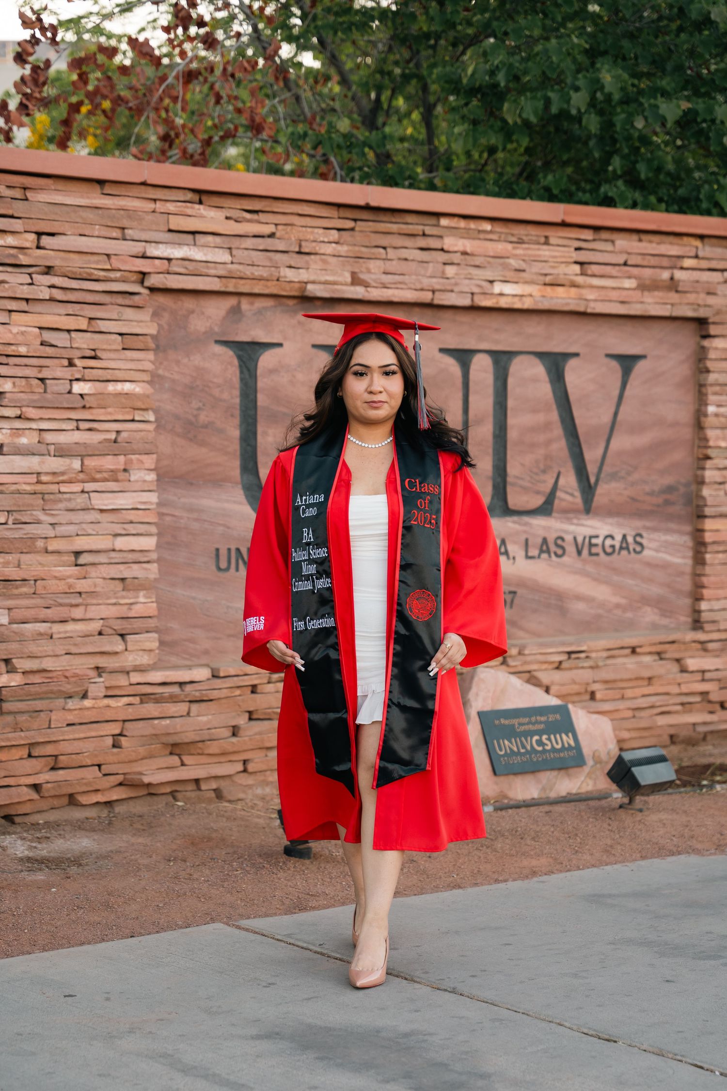 Graduate in red cap and gown poses in front of UNLV stone wall sign.