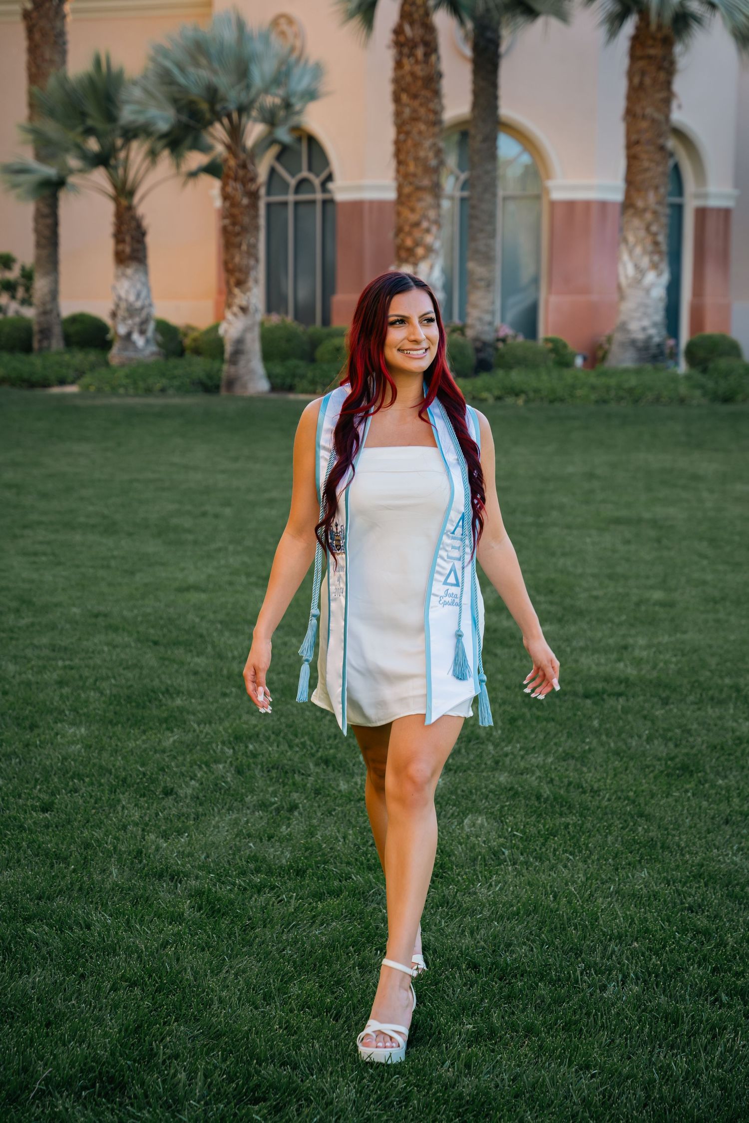 Graduate in white dress and blue stole walks across lawn with palm trees and Spanish architecture.
