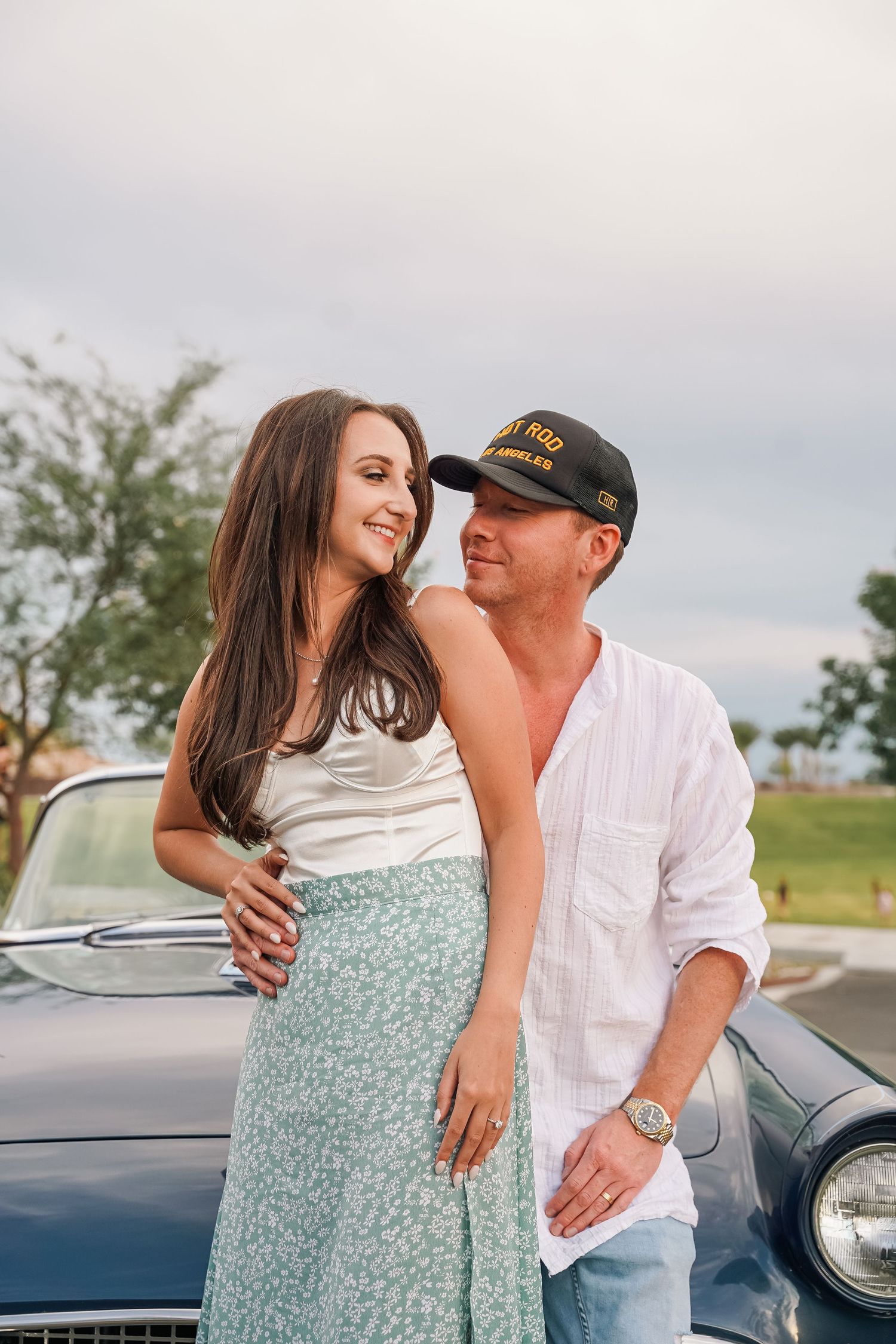 Romantic couple pose by vintage car, wearing casual summer outfits in warm sunlight.