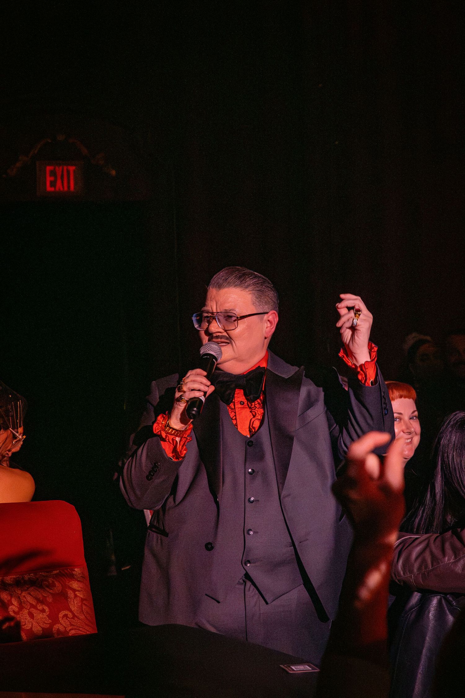 A performer in a dark suit with red bowtie gestures while speaking into a microphone.