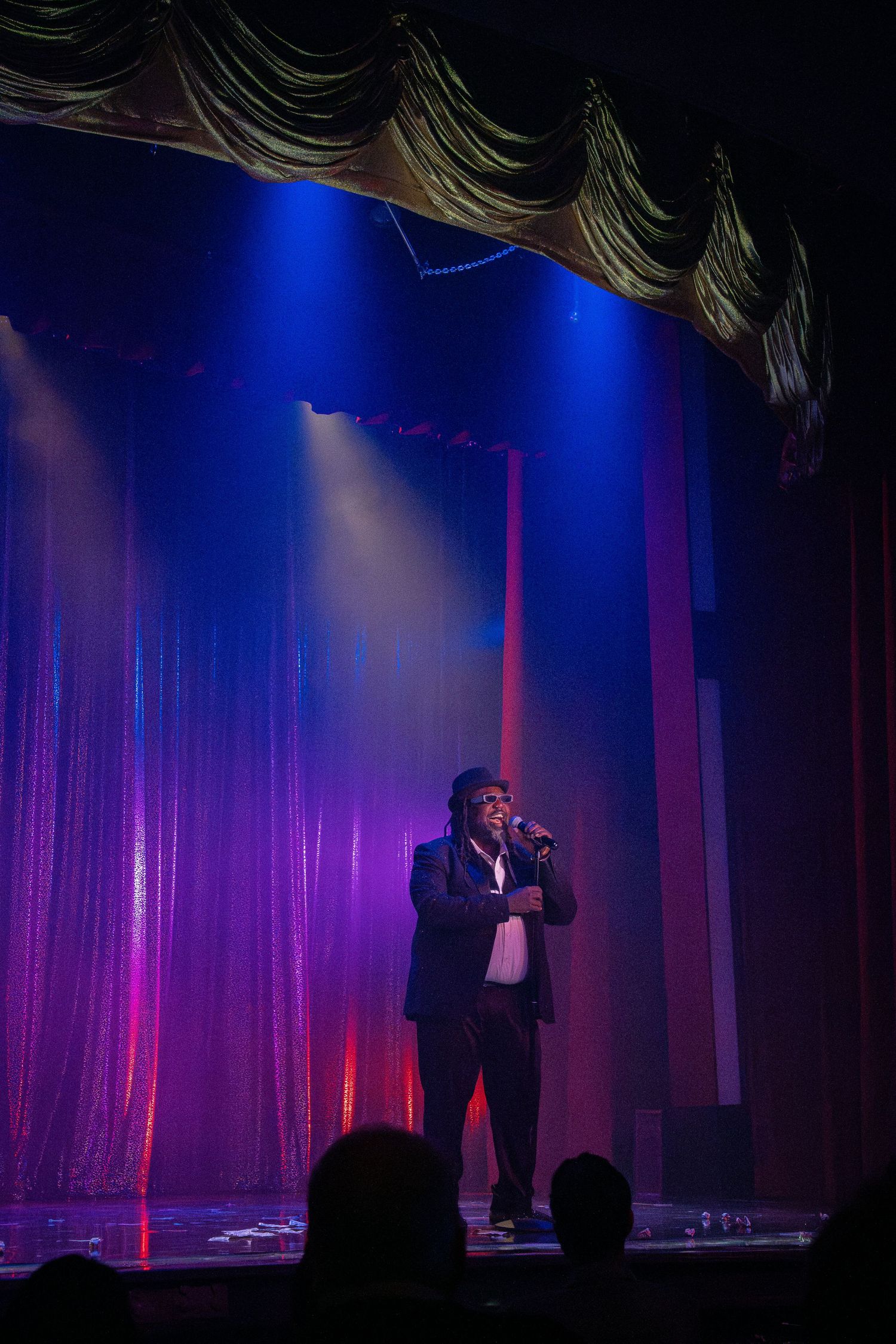 A performer in a suit stands on stage under dramatic blue and purple theatrical lighting.