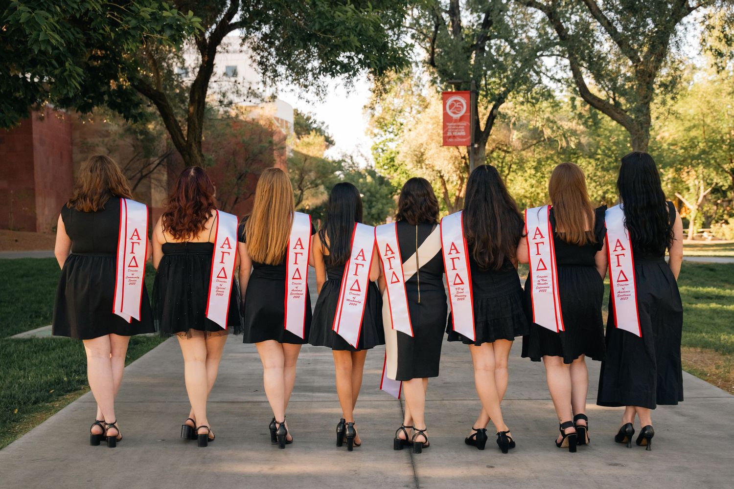 Back view of graduates in black dresses with pink sashes standing in line on campus pathway.