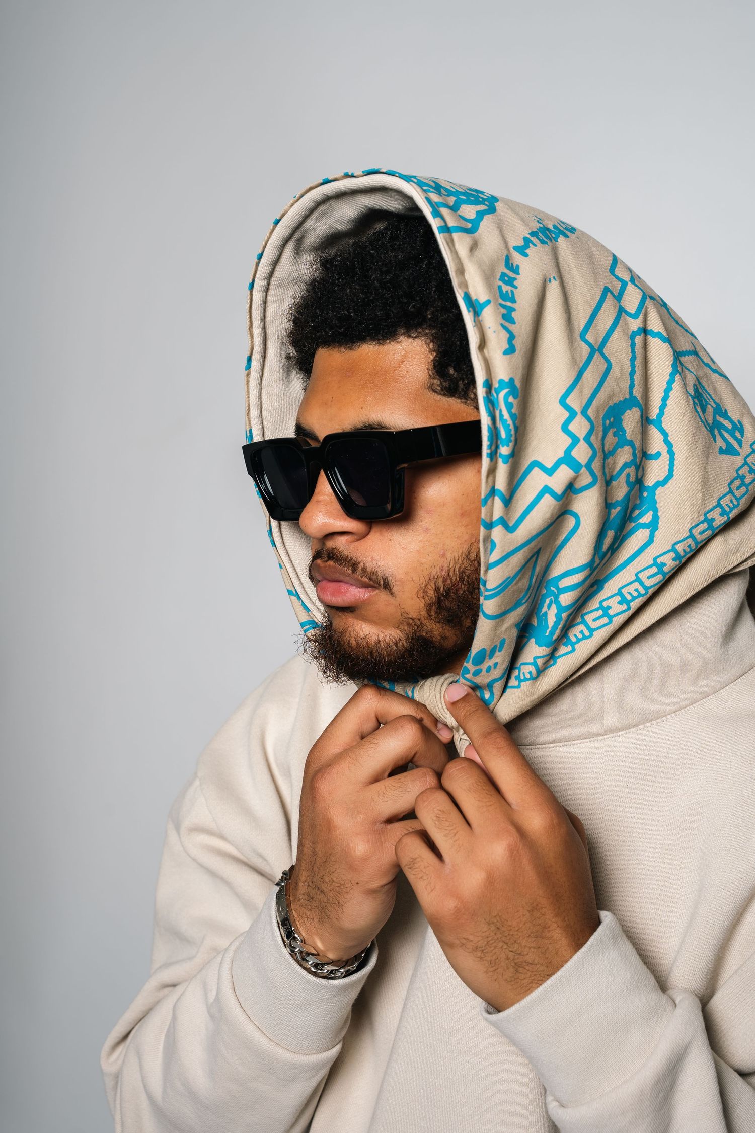 Monaco Media. Portrait of person wearing patterned headscarf, sunglasses and cream hoodie against light background.