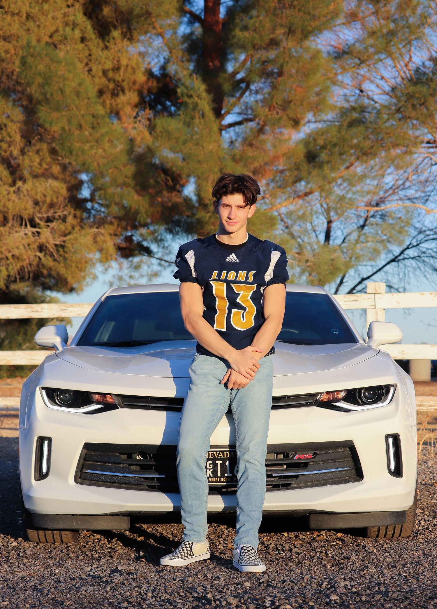 Student athlete in Lions #13 jersey poses in front of white Camaro at sunset.