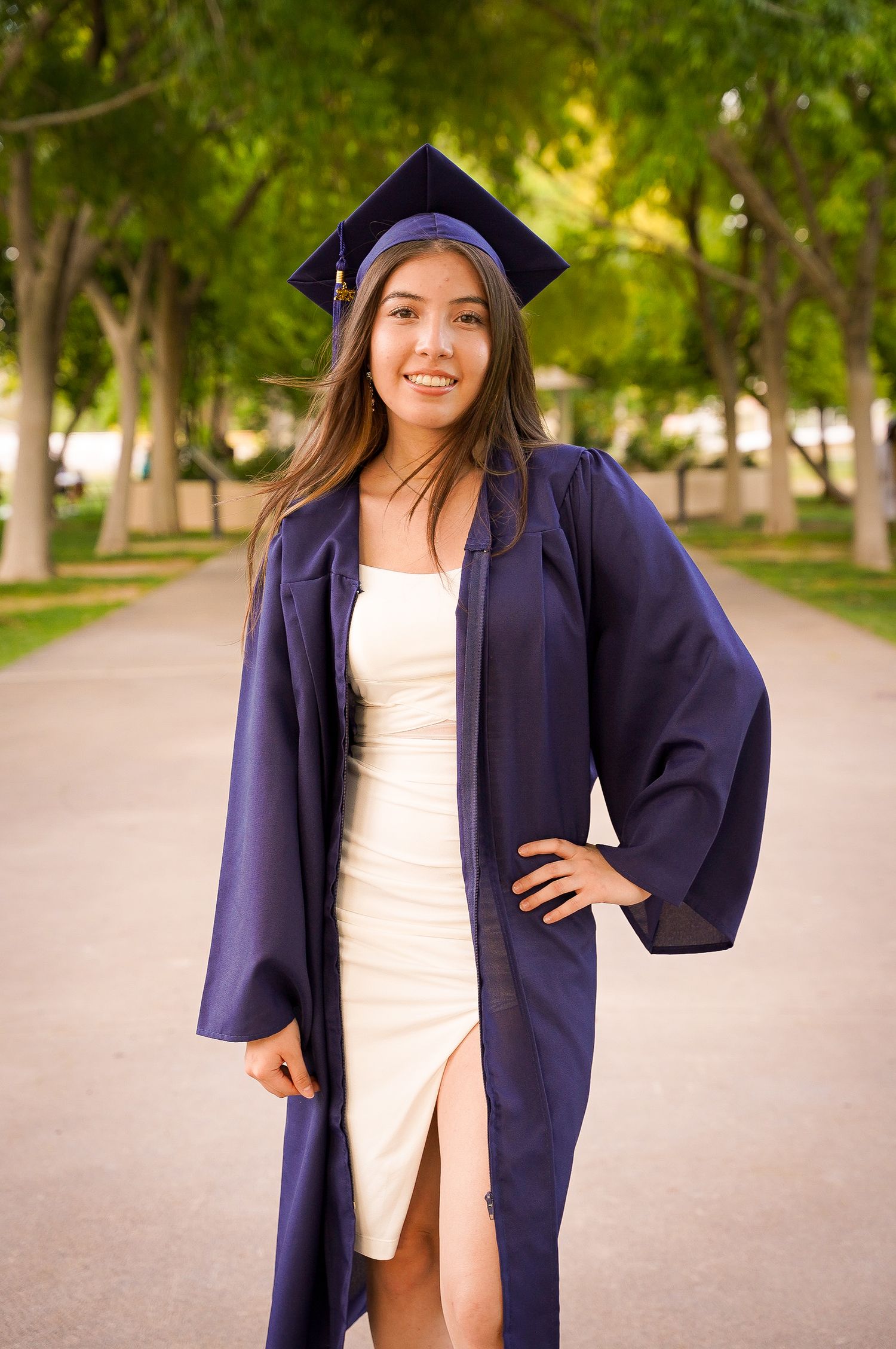 Graduate poses in cap and gown on tree-lined pathway for graduation photos.