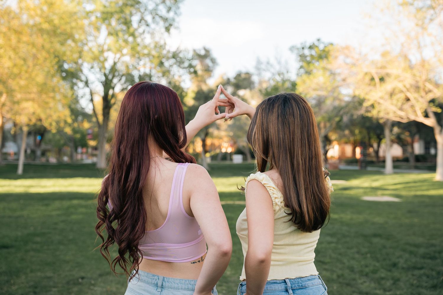 Two friends in crop tops and jeans stand together on a sunny lawn with autumn trees in the background.