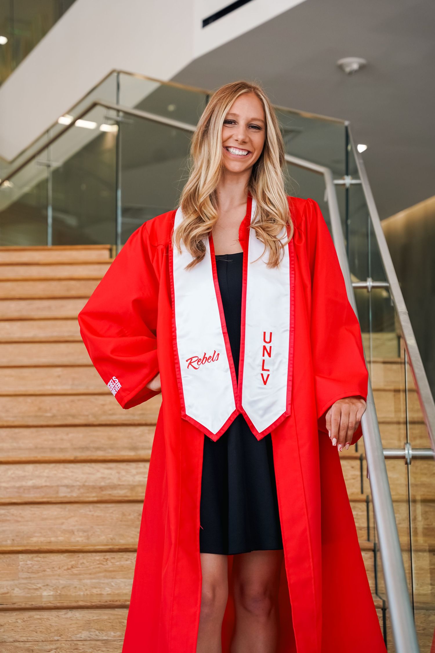 Graduate wearing red academic regalia and white stole standing on wooden steps indoors.