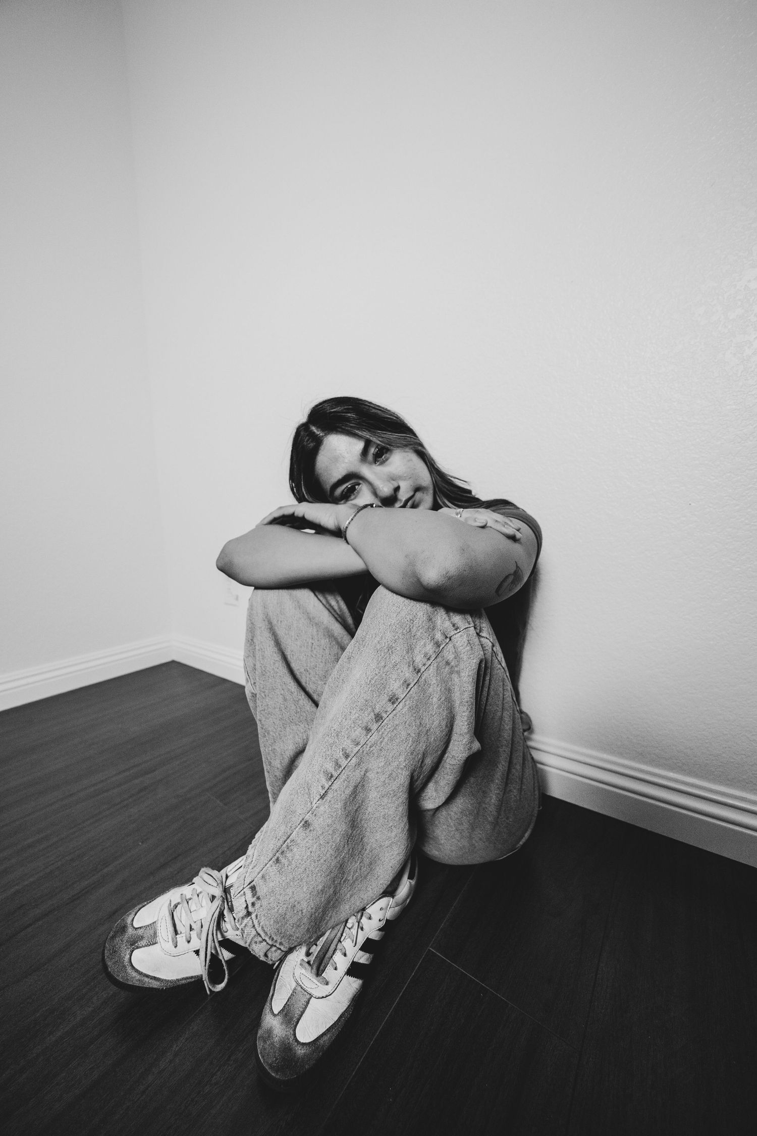 Black and white image of person curled up against a wall wearing jeans and sneakers showing signs of distress or sadness.