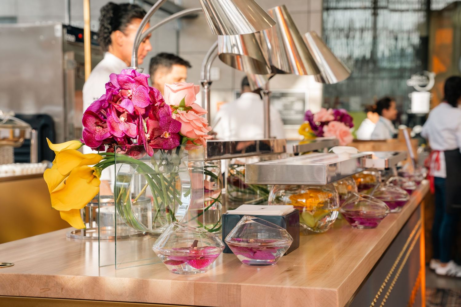 Bright pink flowers in vases decorate a wooden buffet counter with food service items and displays.
