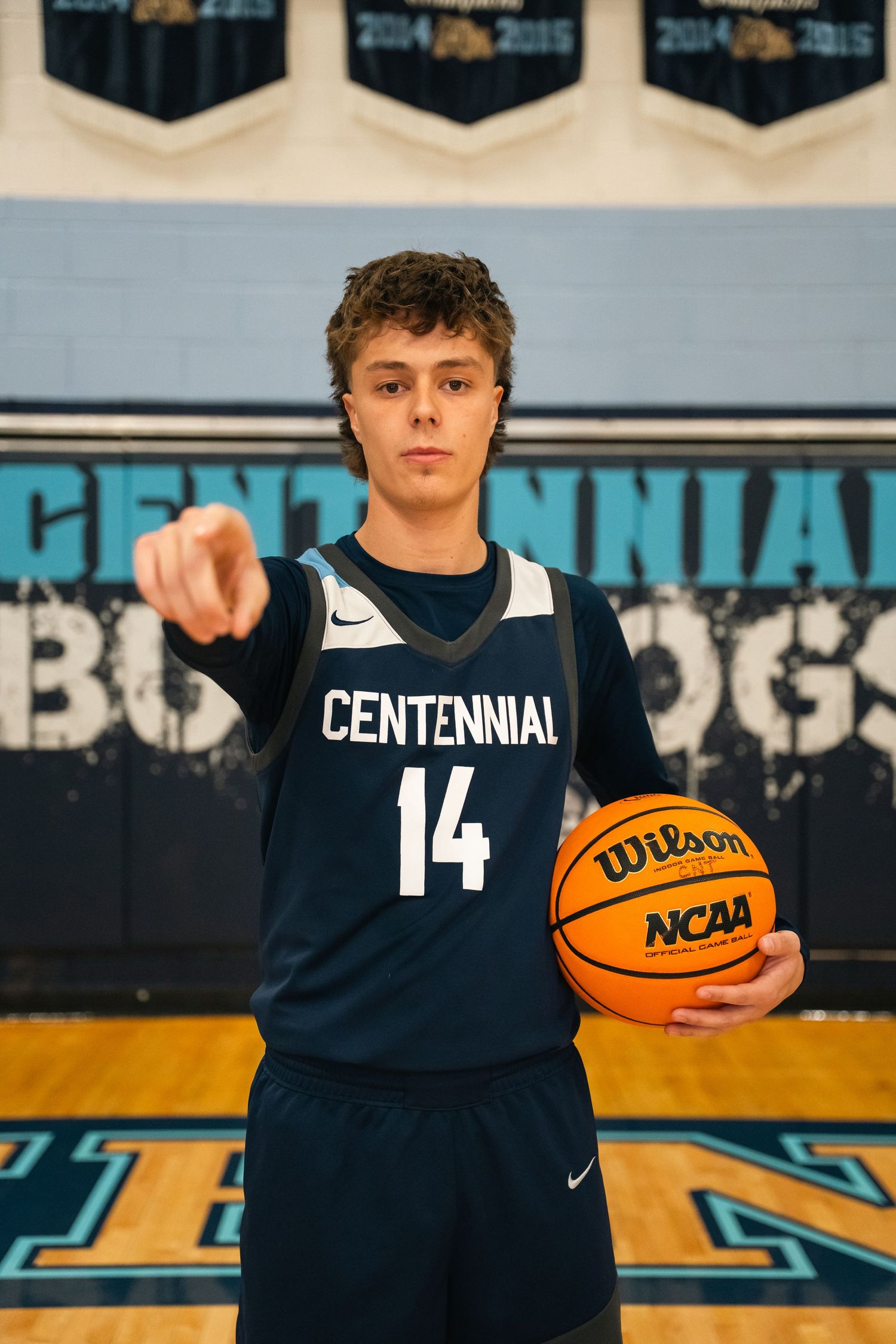 Basketball player in Centennial jersey number 14 pointing while holding NCAA basketball.