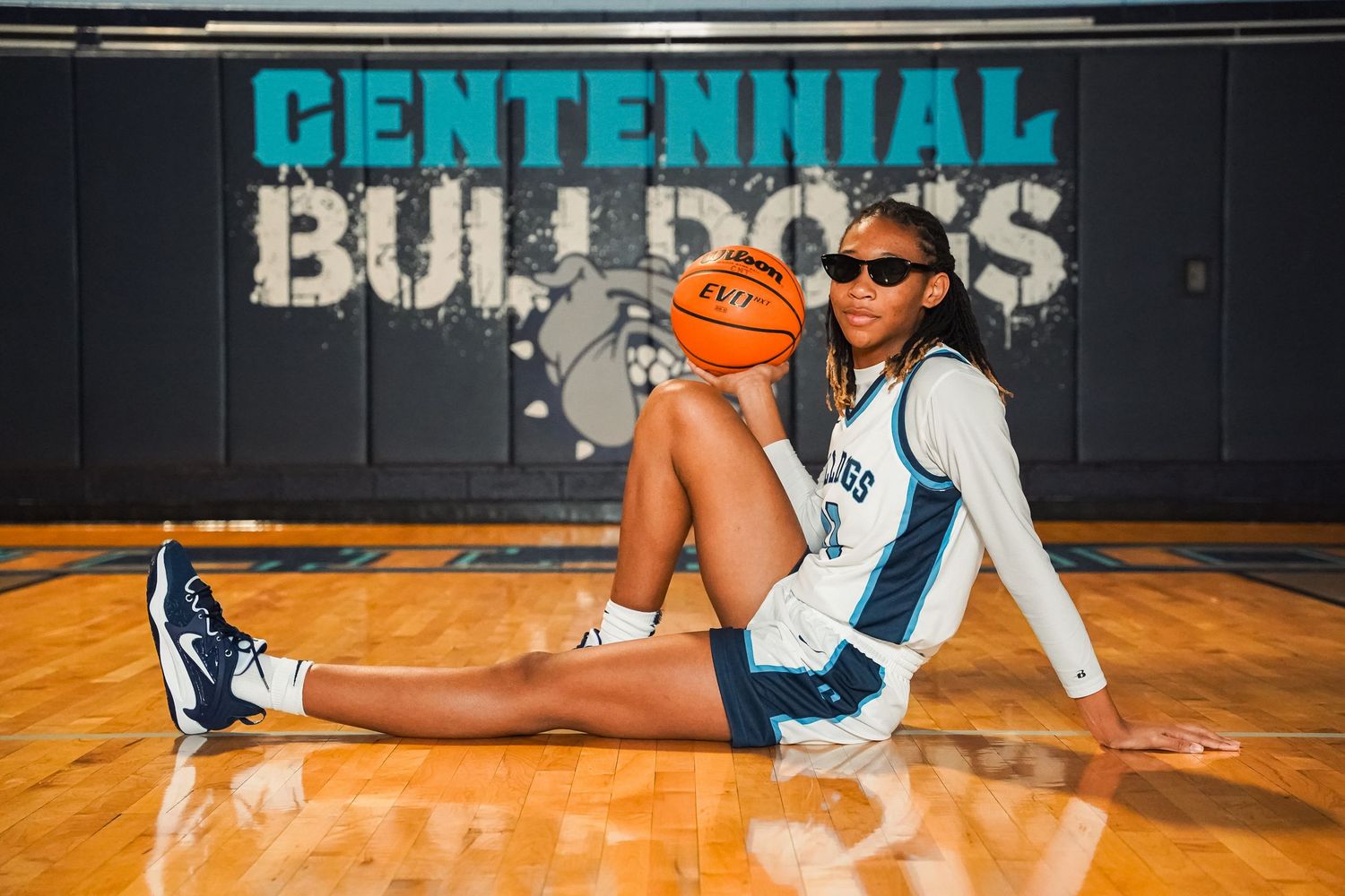 Basketball player in Centennial Bulldogs uniform poses with basketball against gym wall.