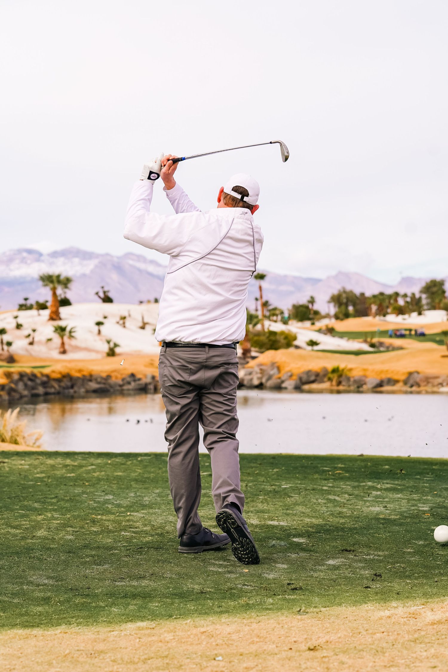 Golfer in white shirt taking a swing near a water hazard on a desert golf course.