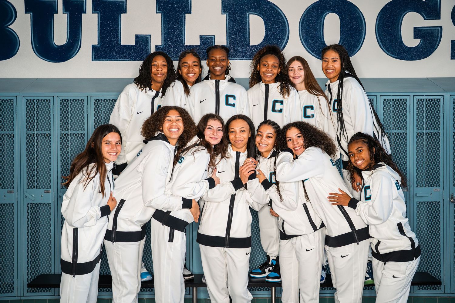 Basketball team wearing white warm-up jackets poses in front of Bulldogs banner.