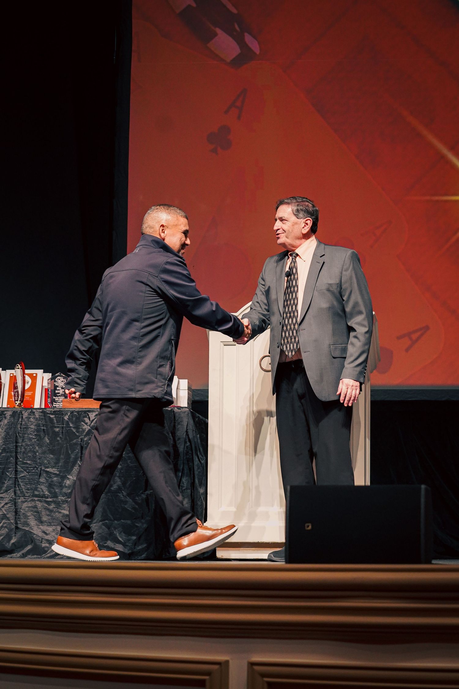 Two people shake hands on stage with playing card backdrop.