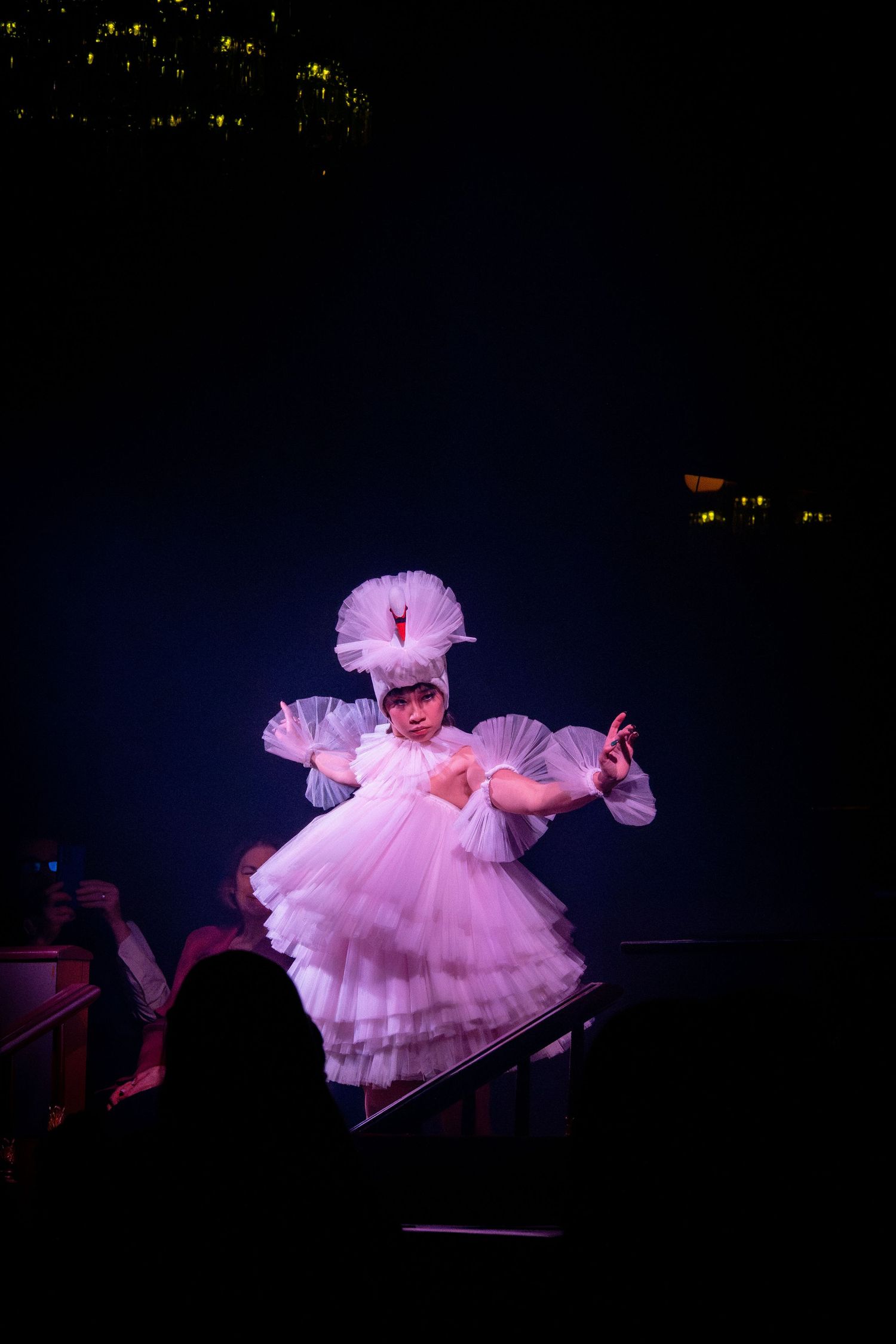 Dancer in layered pink tulle dress performs under spotlight with sparkling effects.