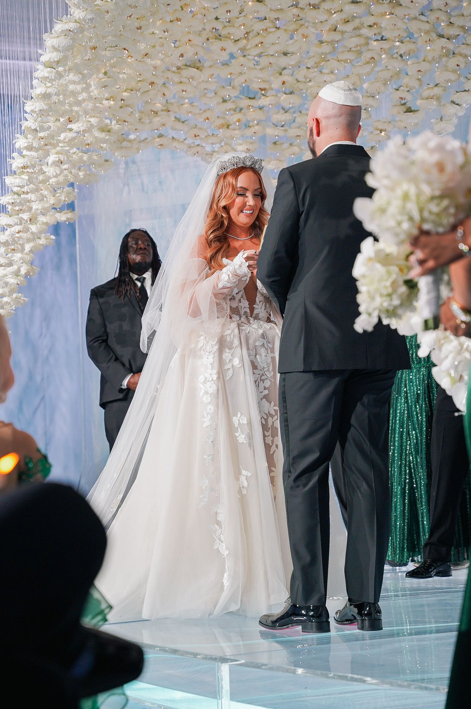 Intimate moment during wedding ceremony under crystal decor with green-dressed attendants.