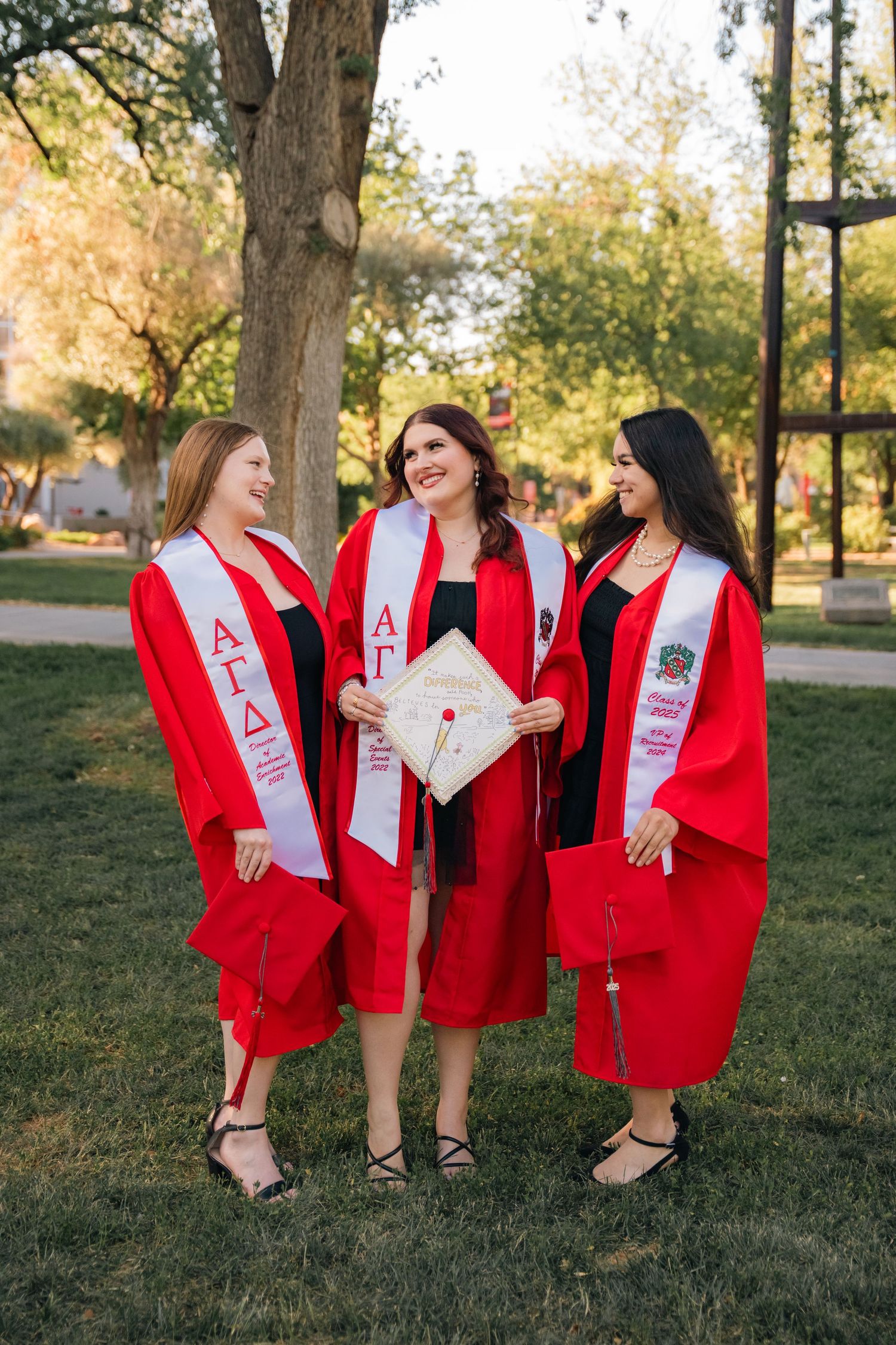 Three graduates in red robes and sashes holding a decorative graduation cap outdoors.