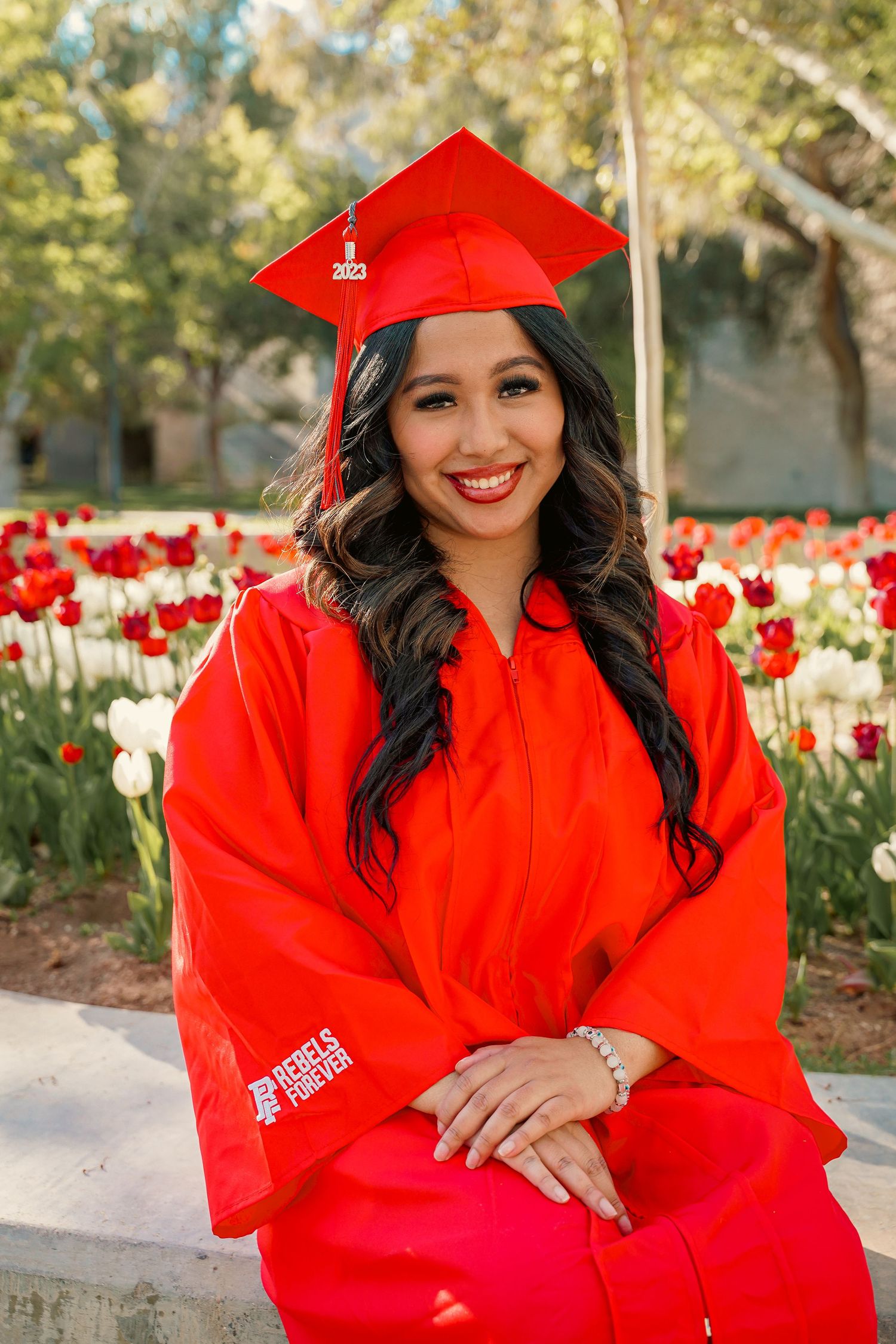 Graduate in red cap and gown poses in front of blooming tulip garden.