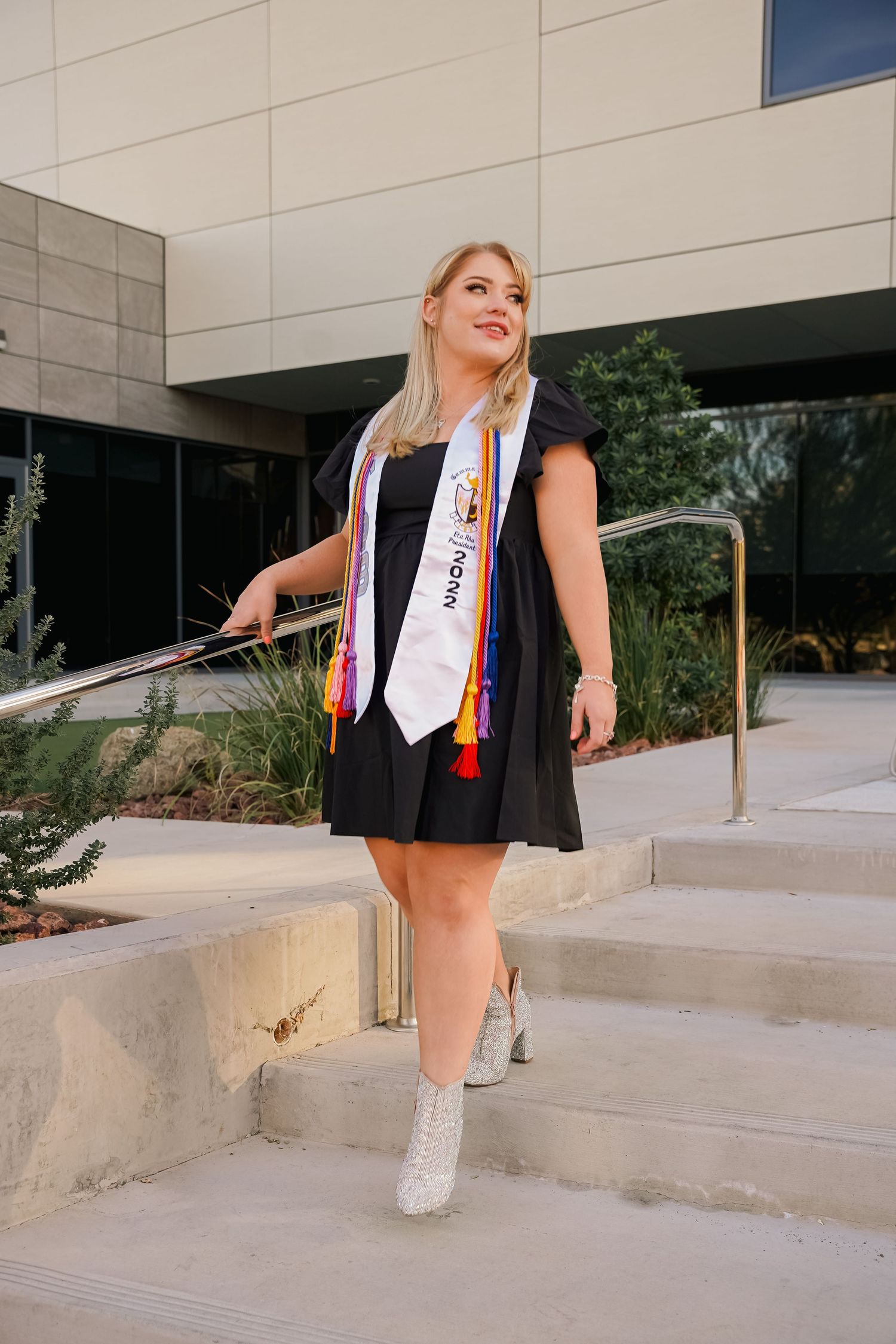 Graduate in black dress with white stoles poses on concrete steps outside modern building.