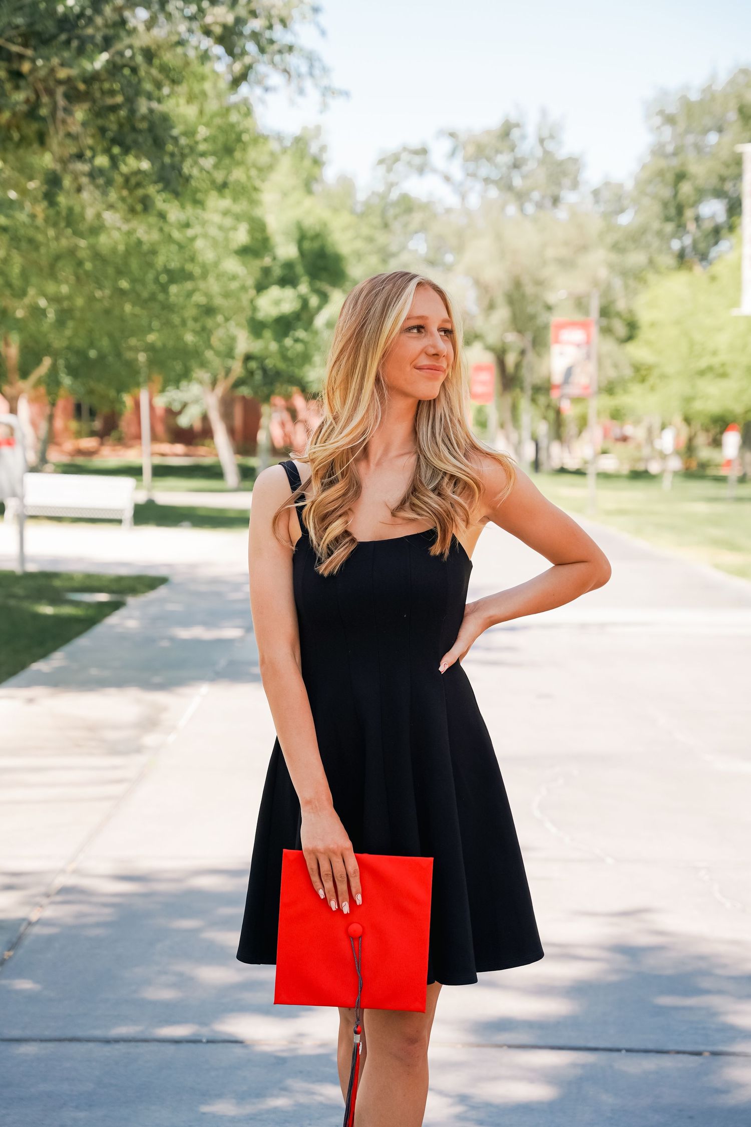 Graduate in black dress holds red diploma cover on tree-lined campus path.