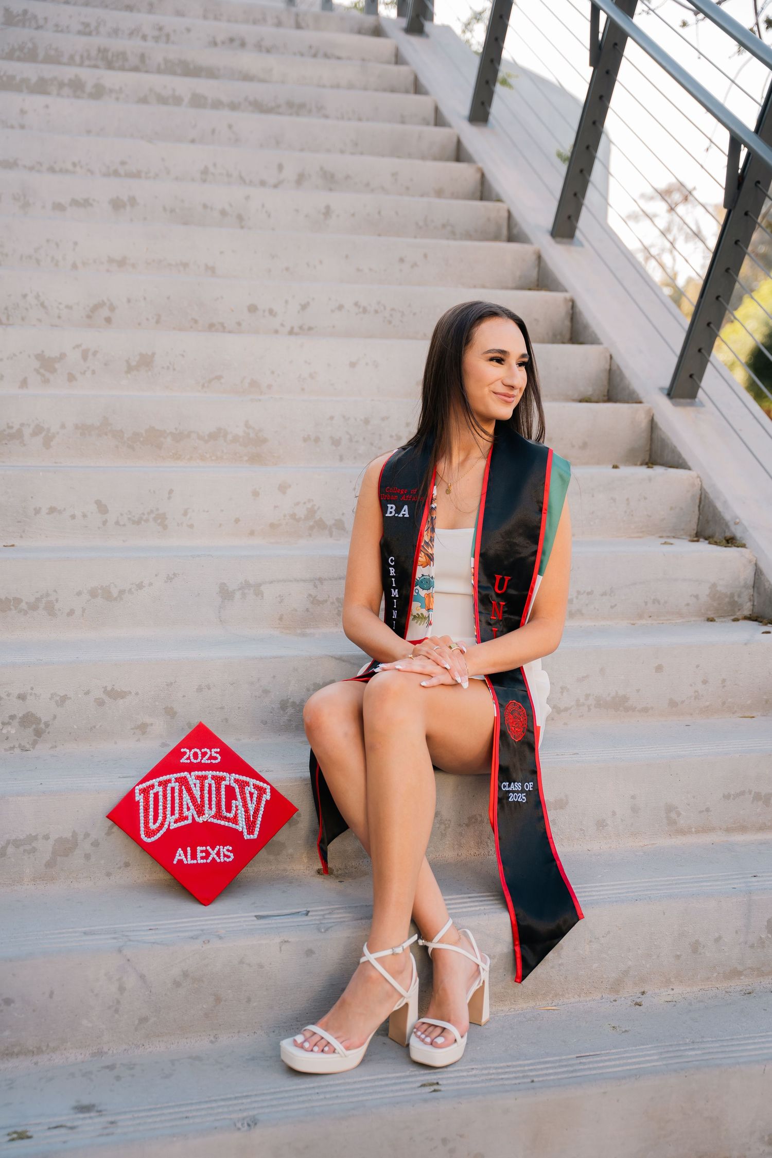 Graduate in white dress with graduation stole sits on concrete steps.