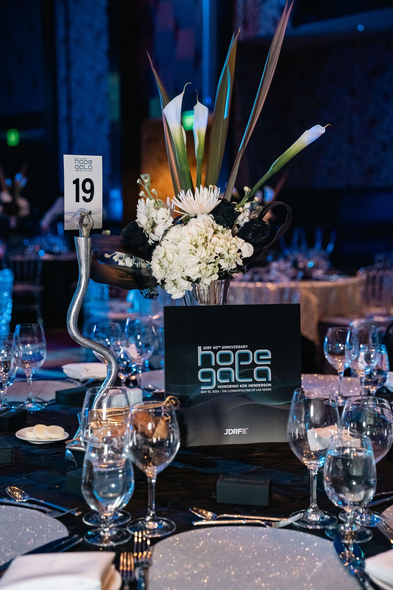 Elegant centerpiece with white flowers and calla lilies on a formal table setting with Hope Gala branding.