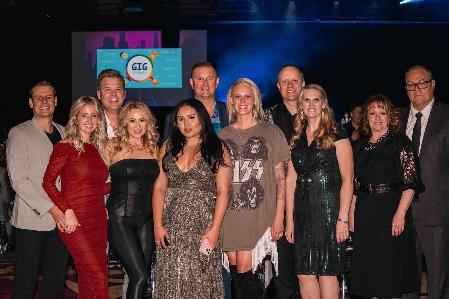 Group photo of formally dressed attendees at a gala event with blue stage lighting.