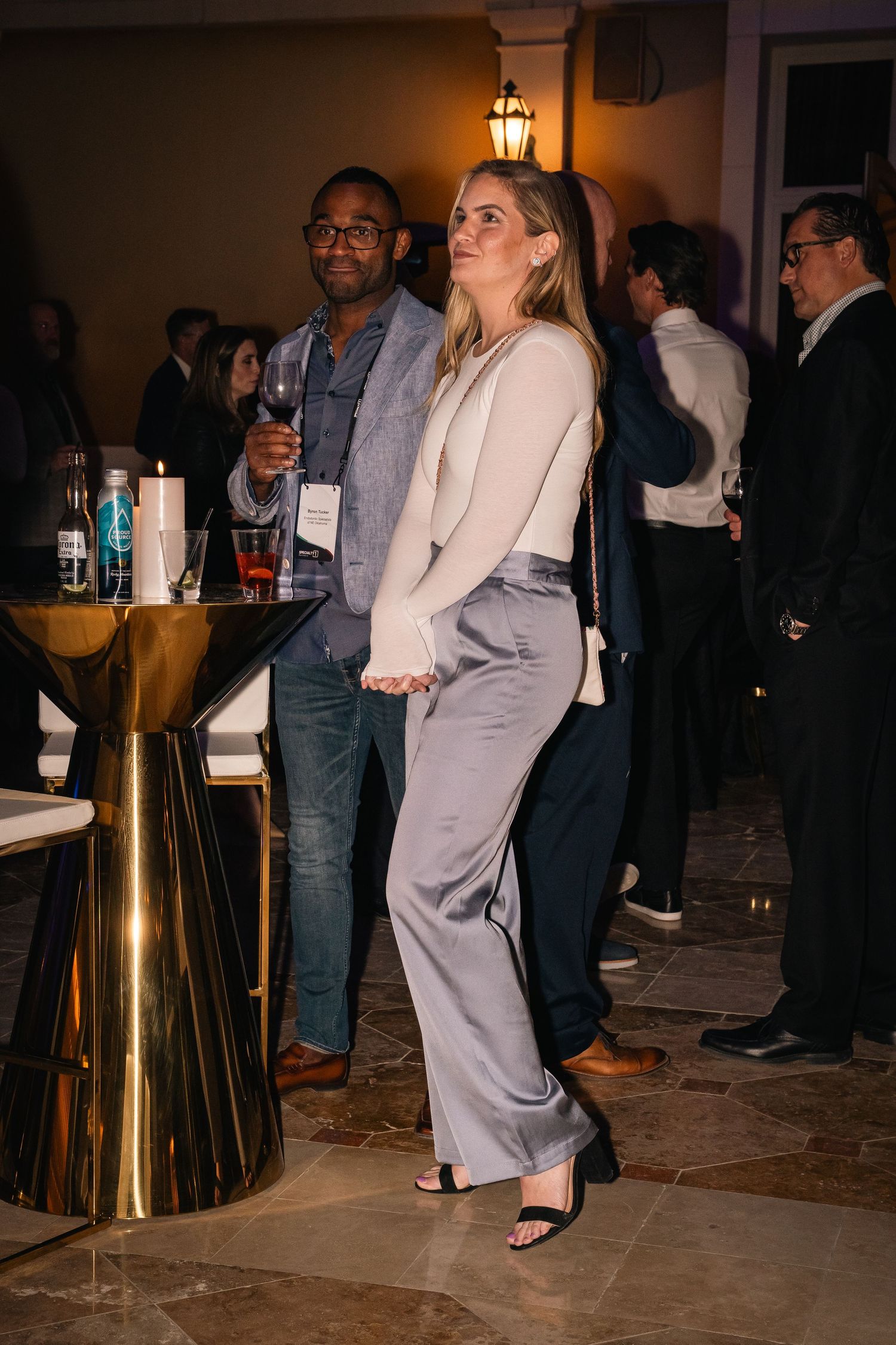Elegantly dressed guests mingle and socialize at an evening networking event near a gold cocktail table.