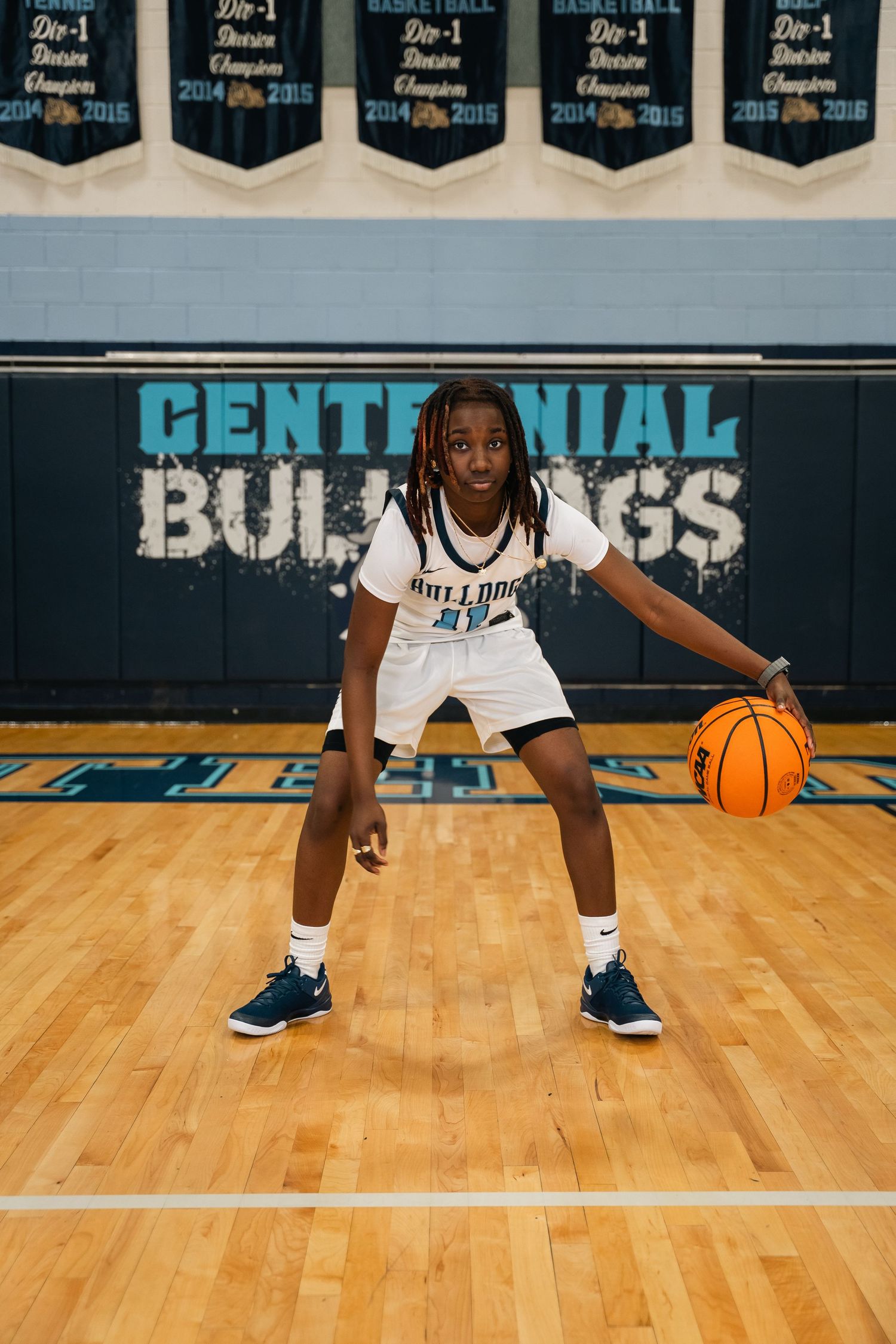Basketball player in defensive stance dribbles ball on wooden court with Centennial Bulldogs logo.