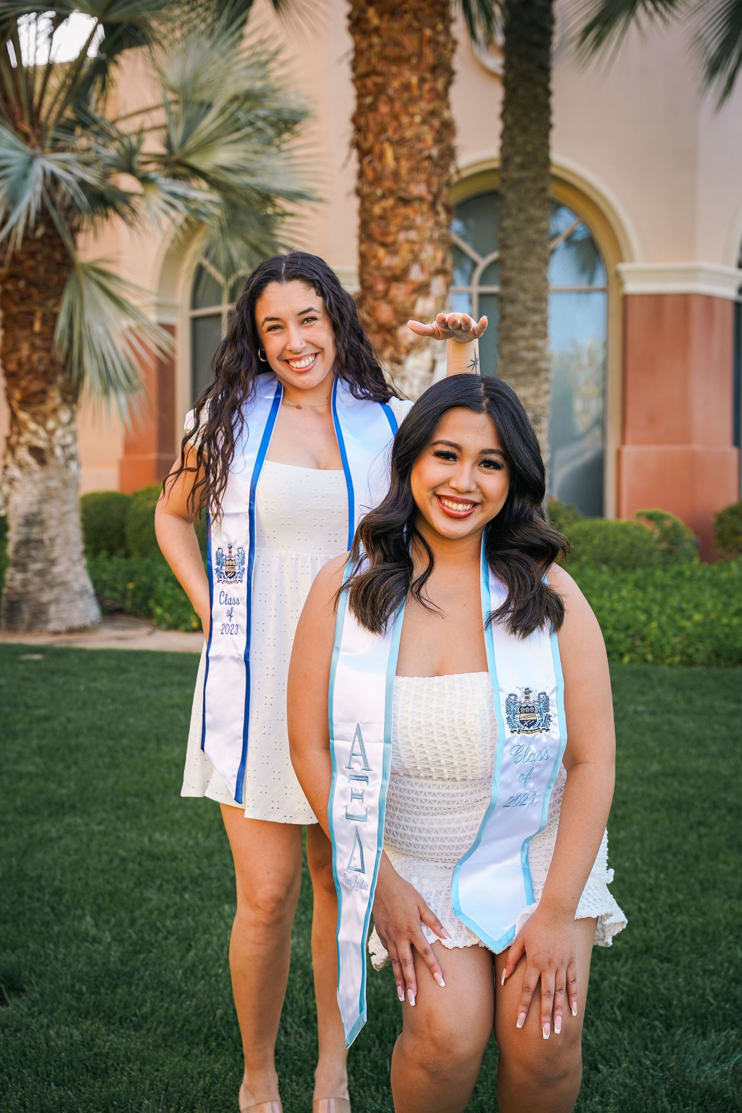 Two graduates wearing white dresses and blue sashes pose for photo in front of palm trees and arched building.
