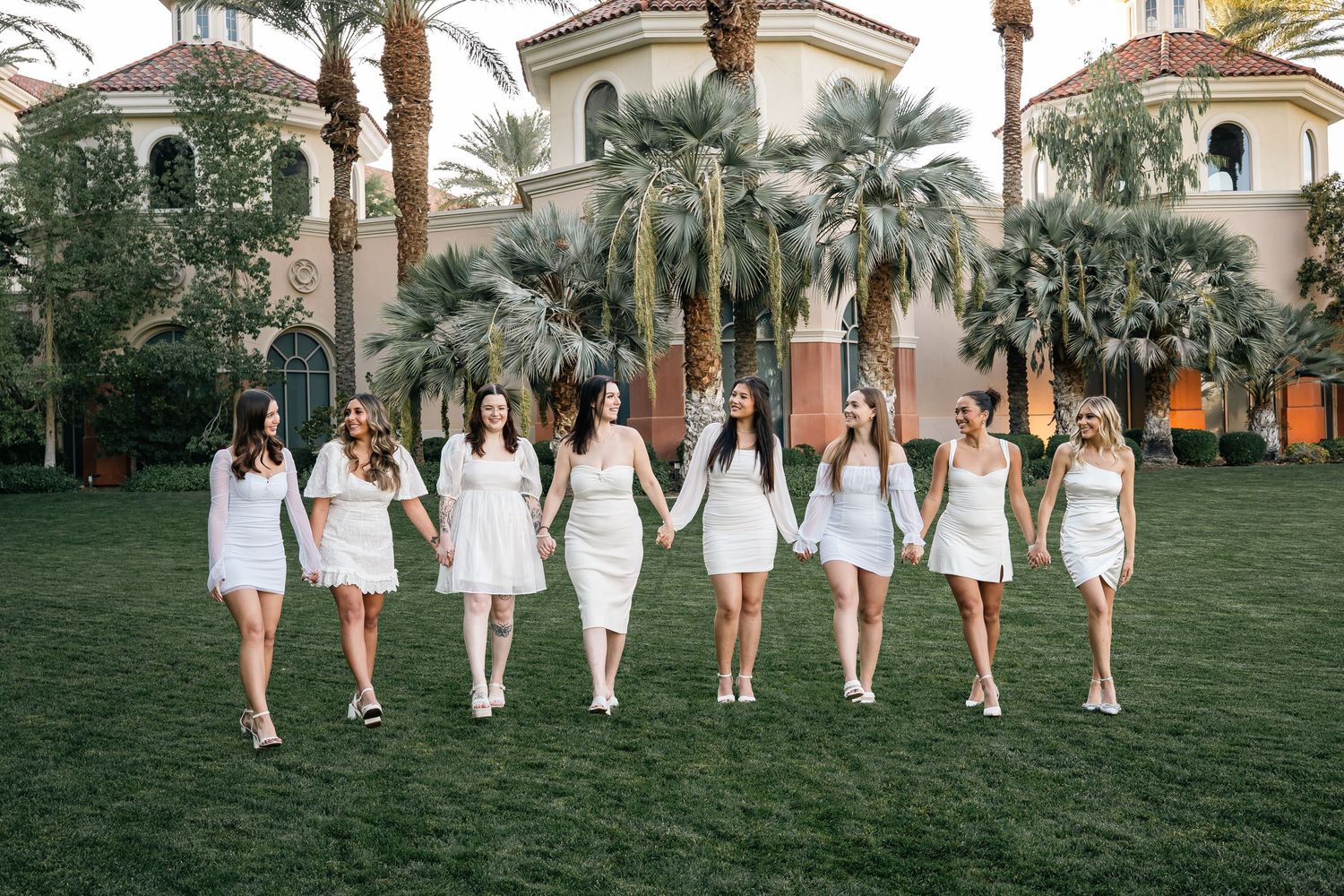 Group in white dresses poses together on grass lawn with Mediterranean-style building and palm trees in background.