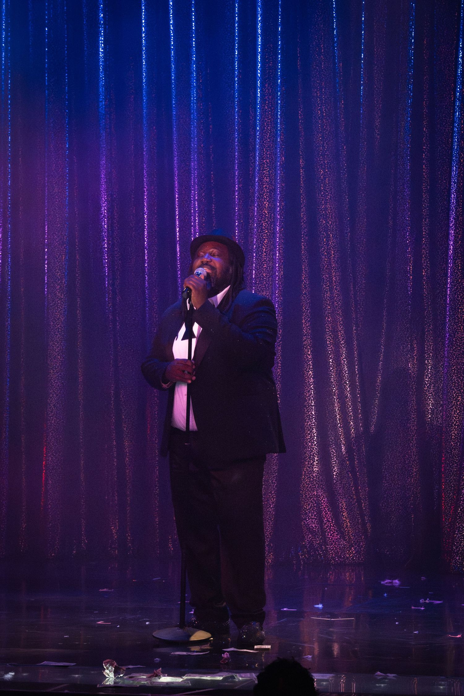A performer in a black suit sings on stage with dramatic blue and purple lighting and sparkly curtains in the background.