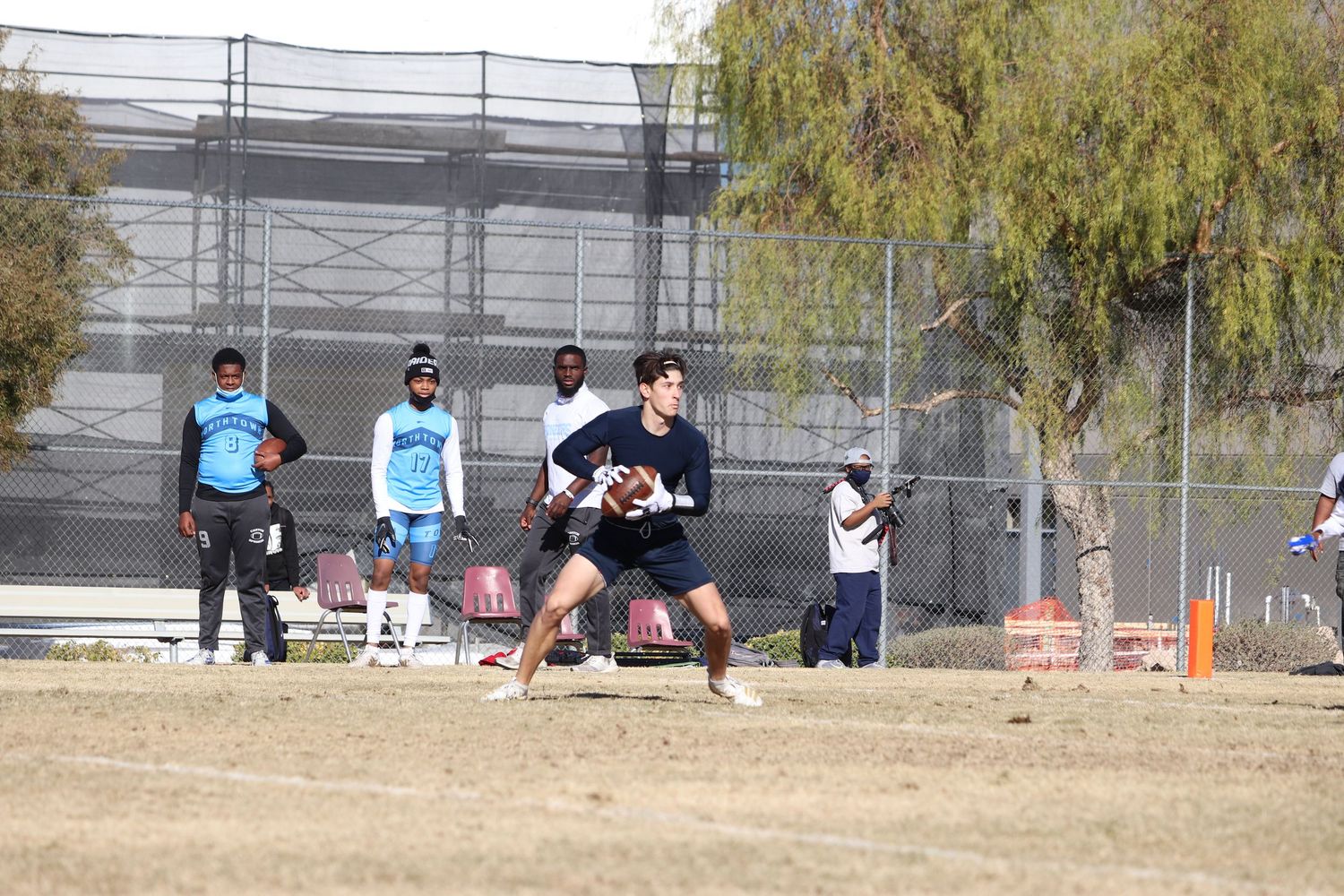 Wide view of sports practice session on dry grass field with chain-link fence in background.