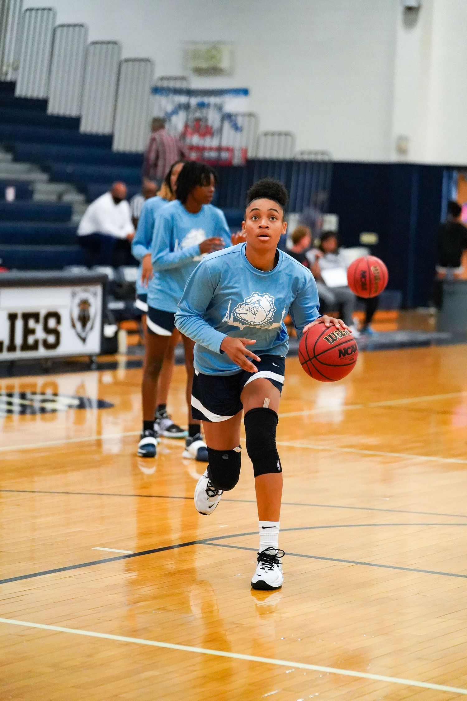 Basketball player in light blue jersey dribbles ball during warmup in gymnasium.