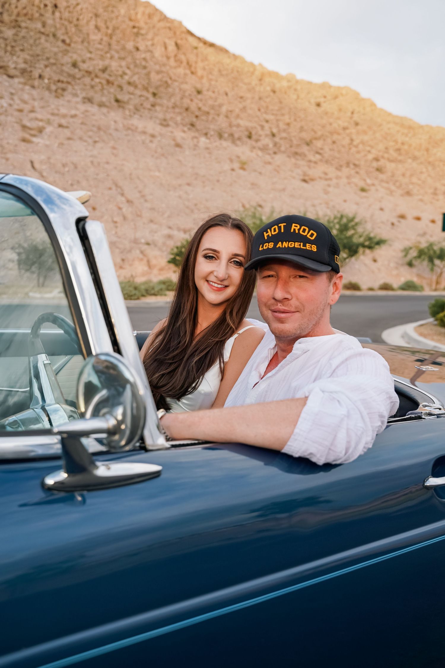 Couple poses with classic convertible car in desert setting during golden hour.