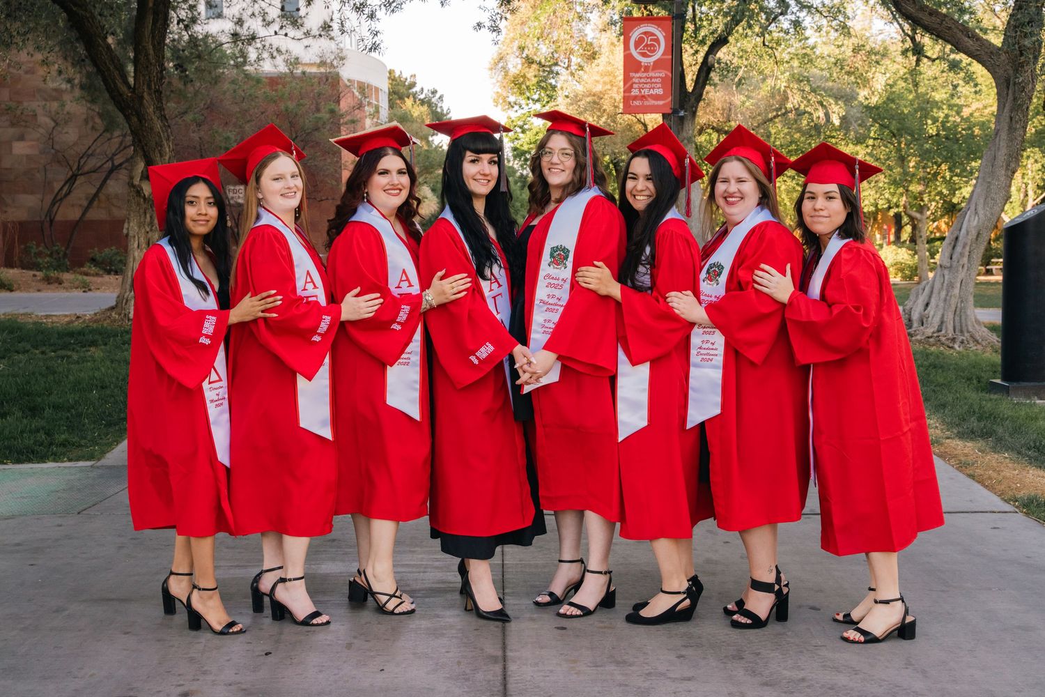 Group photo of graduates in bright red graduation robes and white sashes posing outdoors.