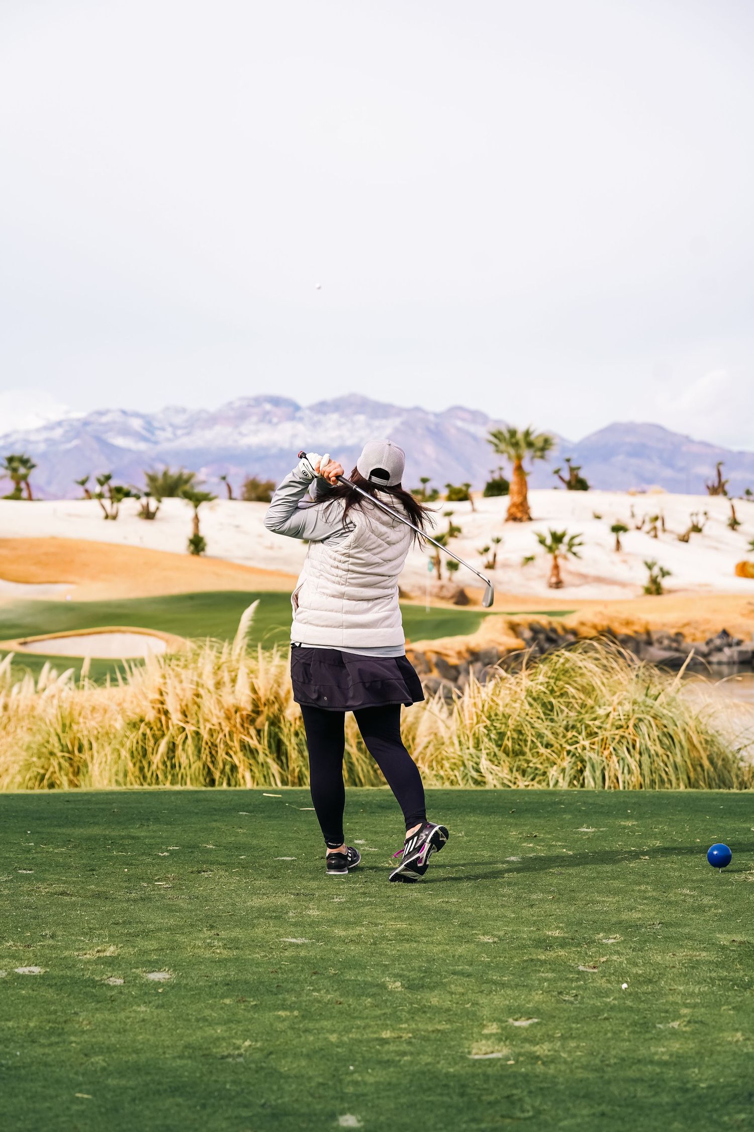 Golfer taking a shot on desert golf course with mountains and palm trees in background.