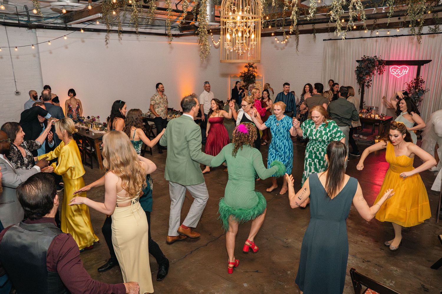 Large group of wedding guests dancing together on industrial venue dance floor with string lights above.