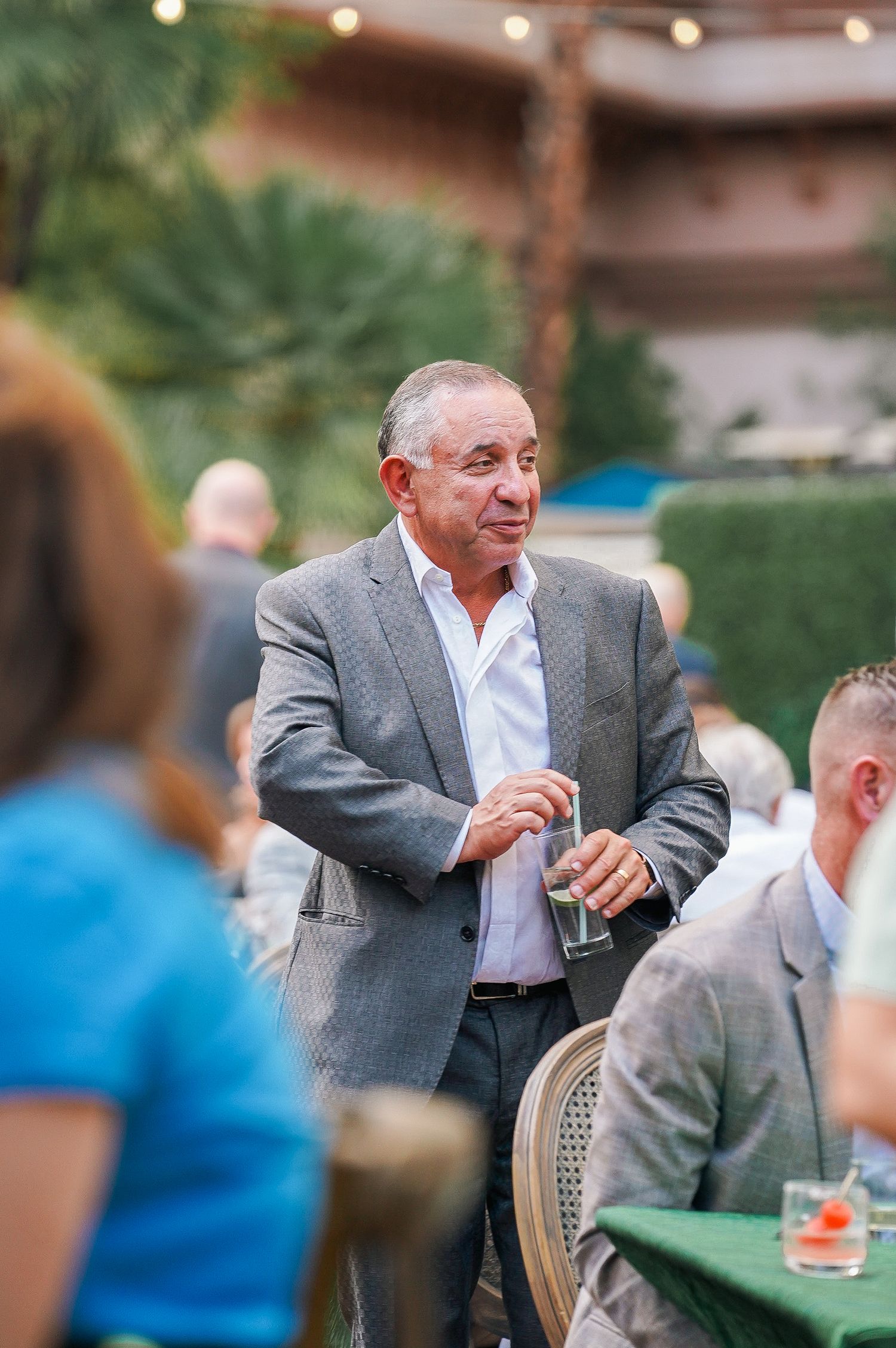 Man in grey suit jacket enjoying drinks and conversation at outdoor gathering.