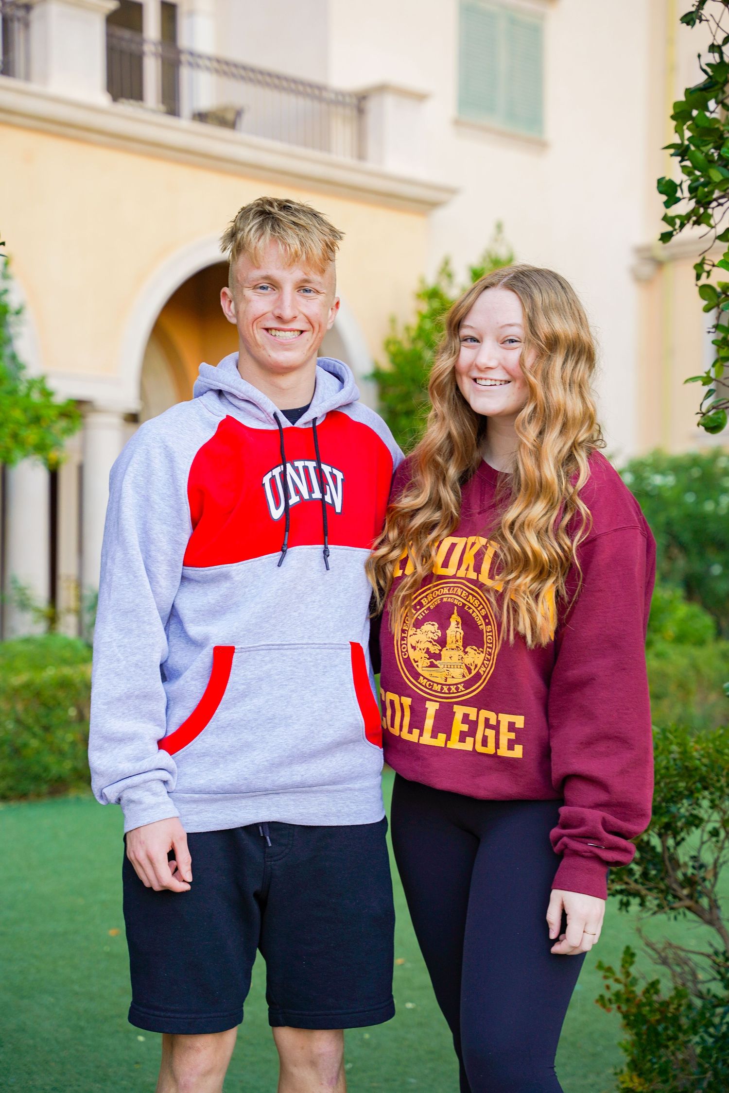 Two students wearing university branded sweatshirts pose together in front of a Mediterranean-style building.