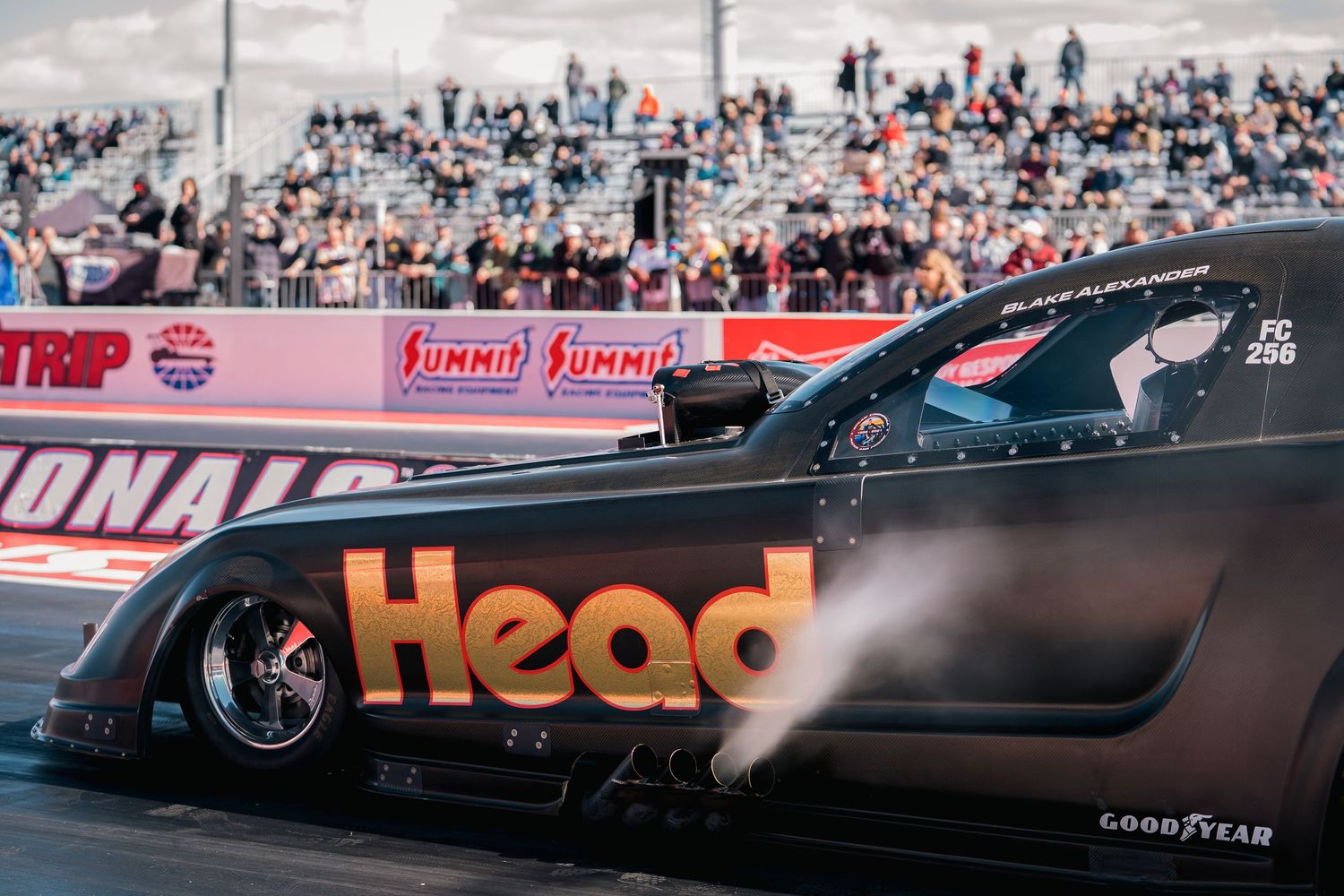 Black drag racing car with 'Head' branding competing on the race track with spectators in background.