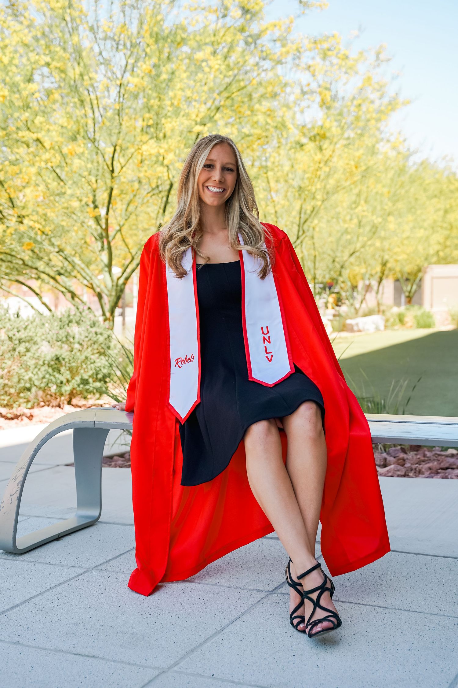 Graduate in red academic regalia relaxes on outdoor chair against desert landscape.