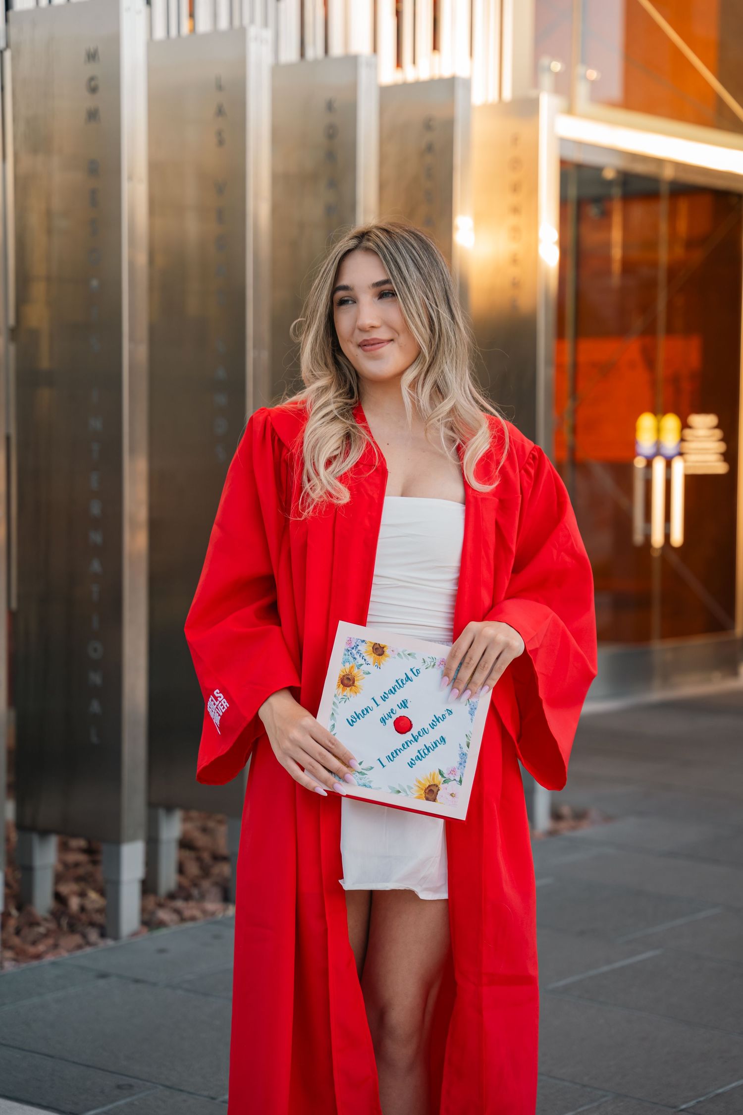 Graduate in red graduation gown poses with decorated cap against metallic backdrop.