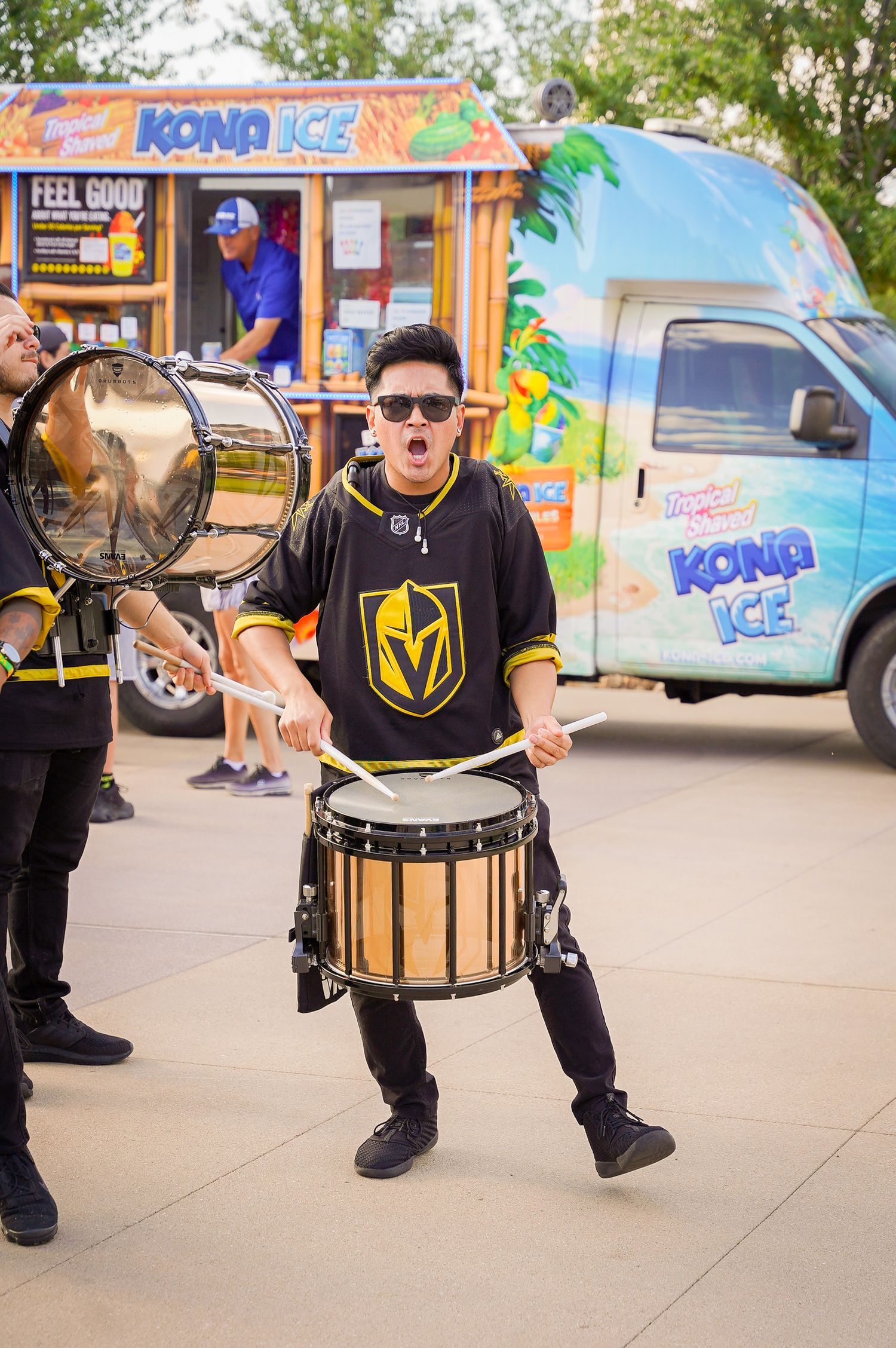 Drummer performs in Vegas Golden Knights jersey near Kona Ice truck at outdoor event.