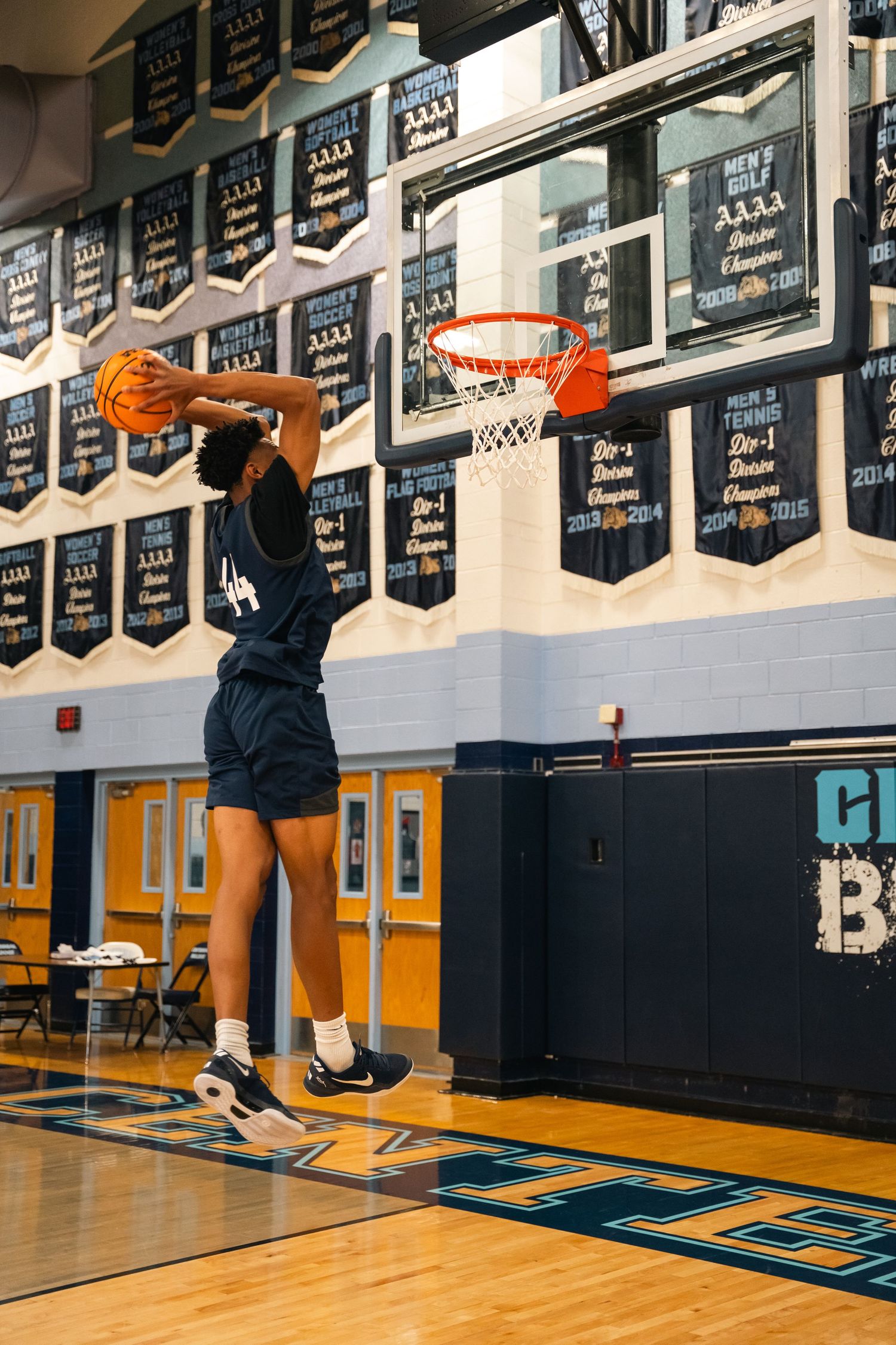 Player in navy uniform jumping for a slam dunk against wall of championship banners.