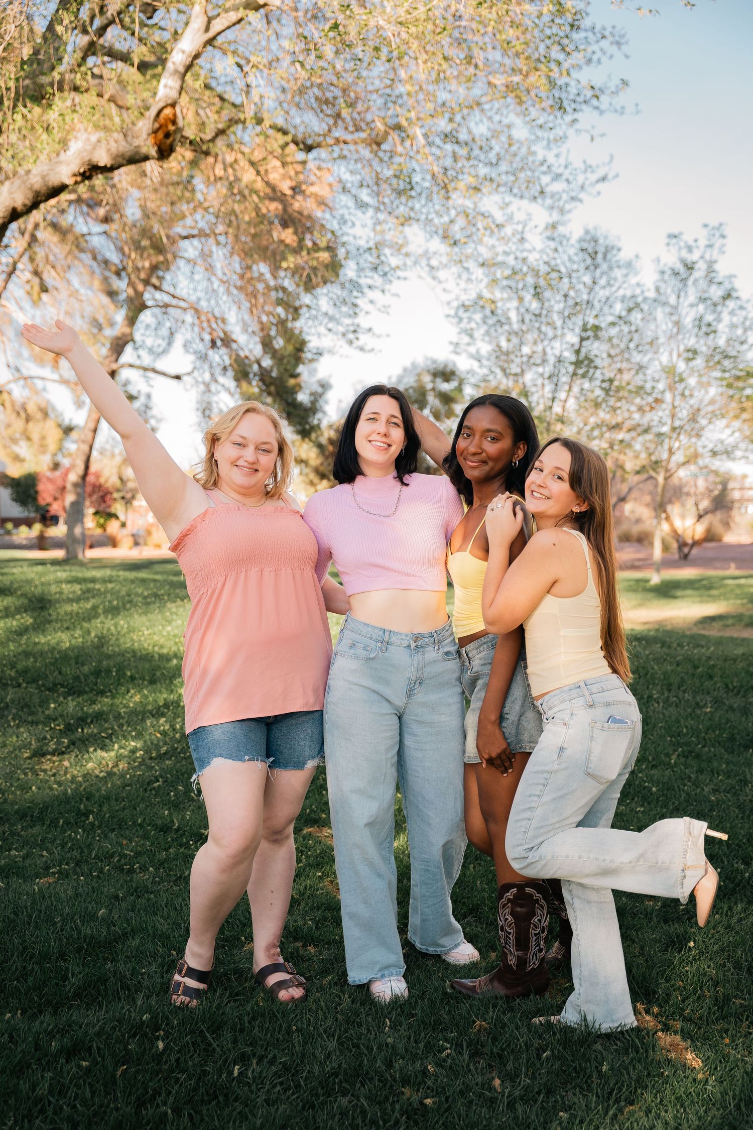 Four friends pose together on a sunny lawn wearing casual spring outfits in pastel colors.
