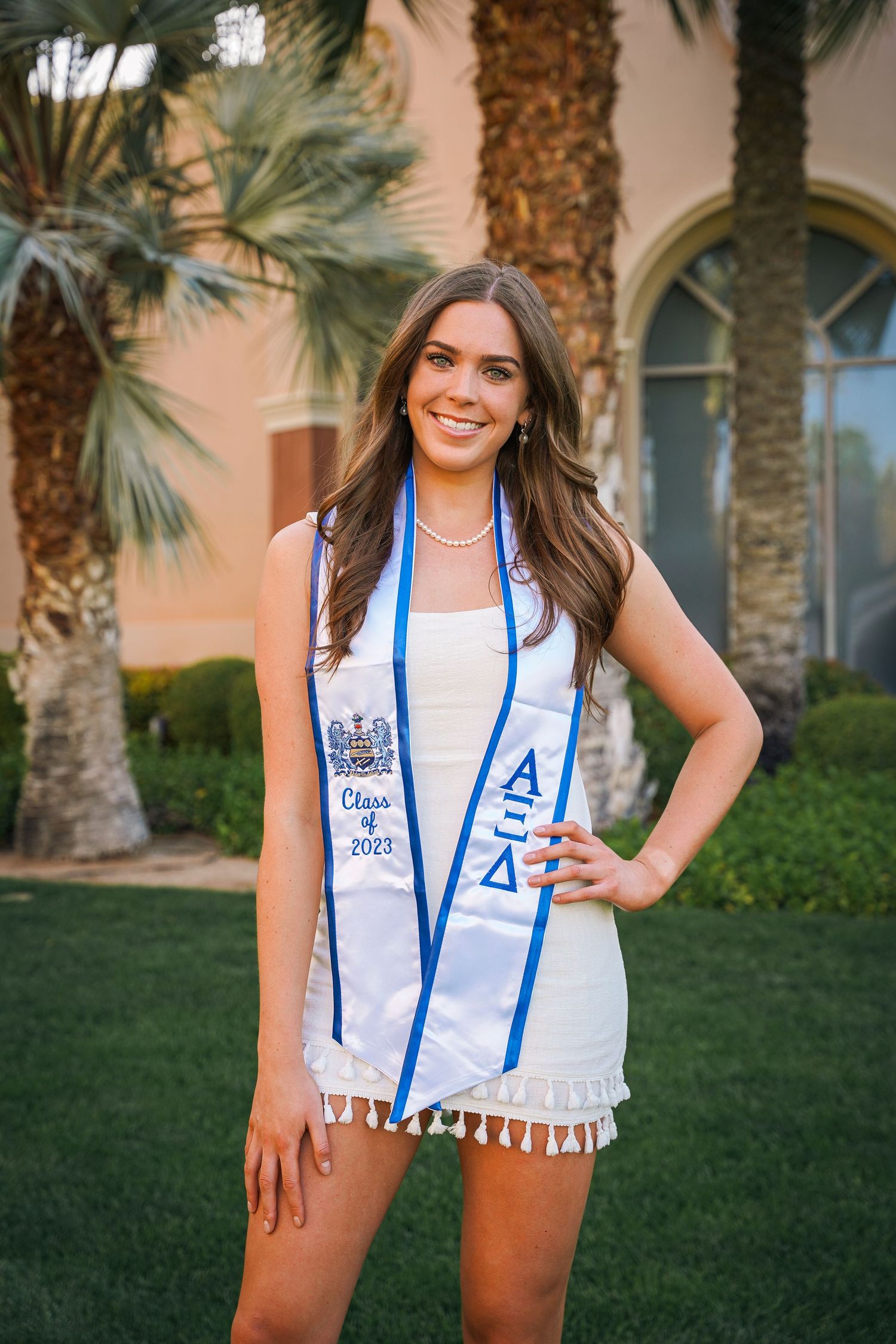 Graduate in white dress and blue sash poses on lawn with palm trees and Mediterranean architecture behind.