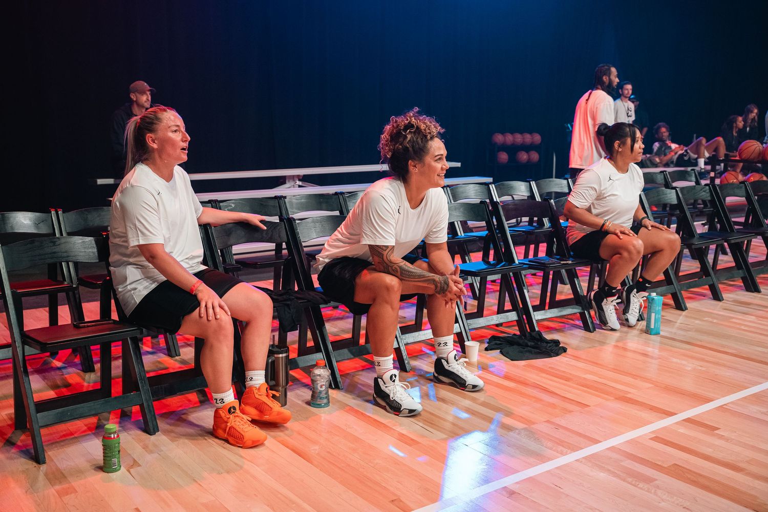 Row of players sitting on chairs along sideline of indoor basketball court.