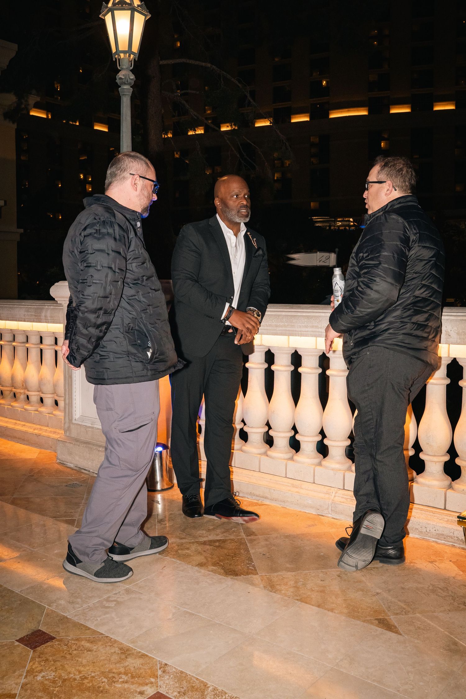Three guests engaged in networking conversation on outdoor terrace with ornate railing and lamp post.