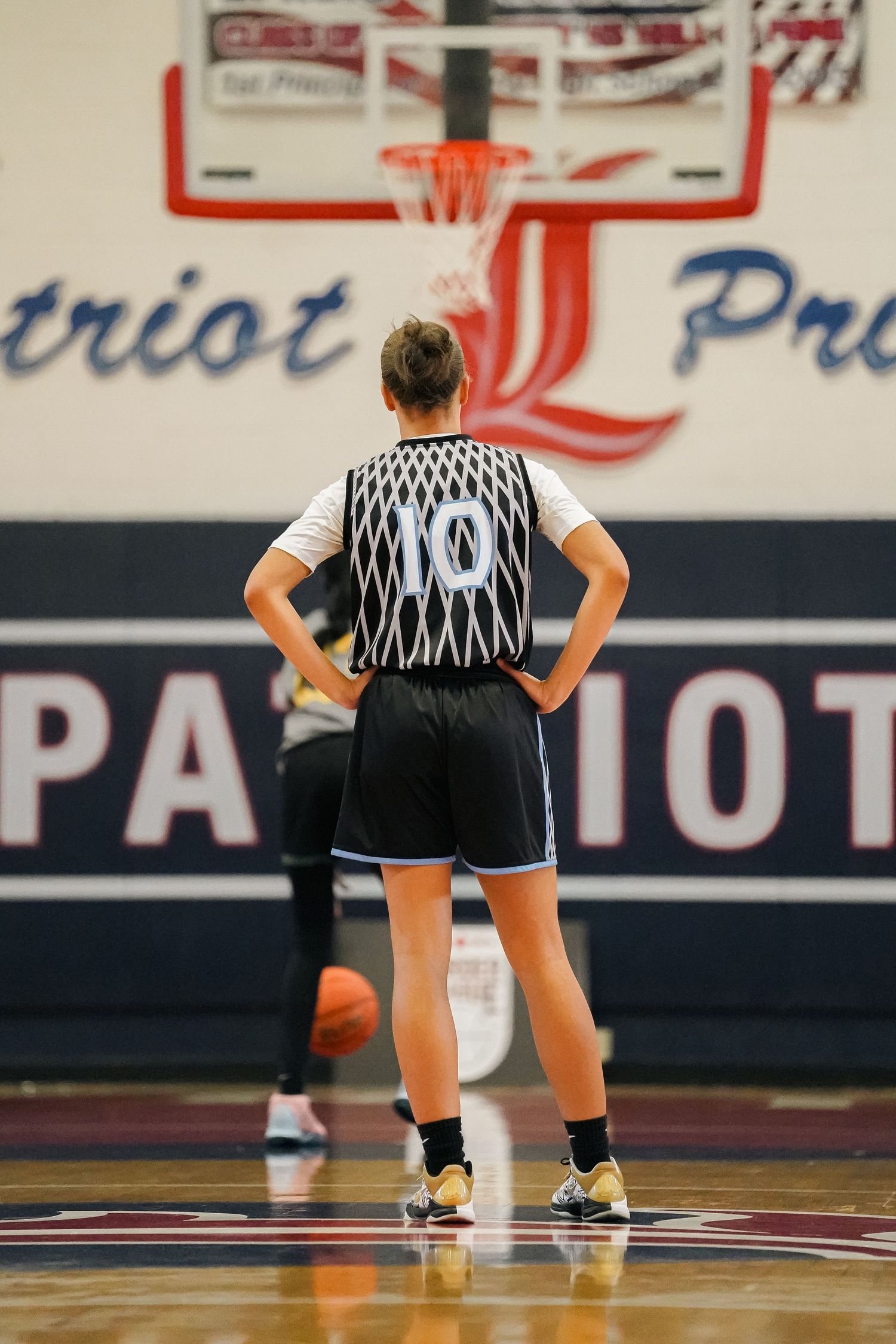 Basketball referee stands on court with hands on hips in front of Patriot sign.